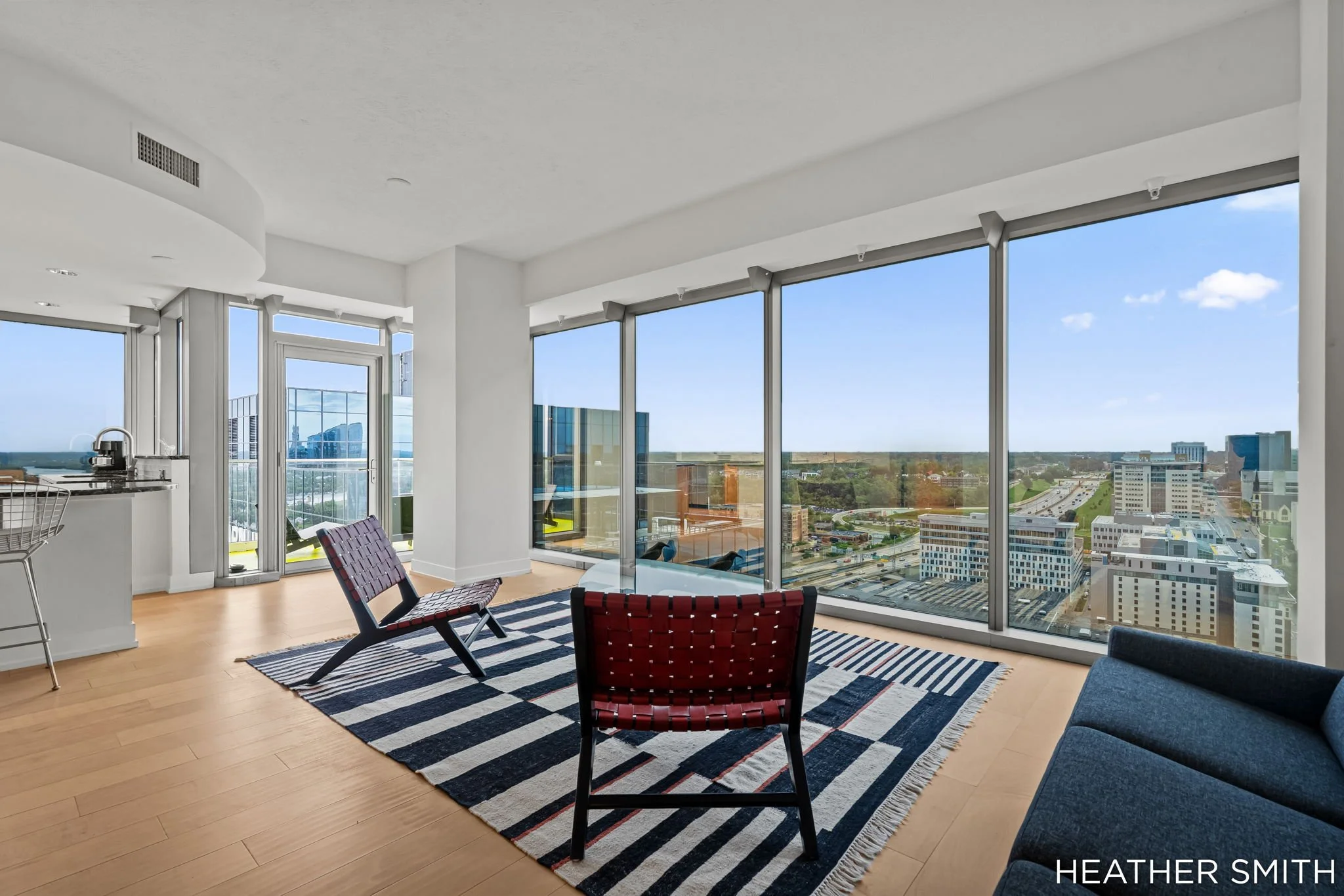 Interior view of a modern high-rise apartment with large floor-to-ceiling windows showing a cityscape and clear blue sky, furnished with a dark blue armchair, a red woven chair, and a black and white striped rug.