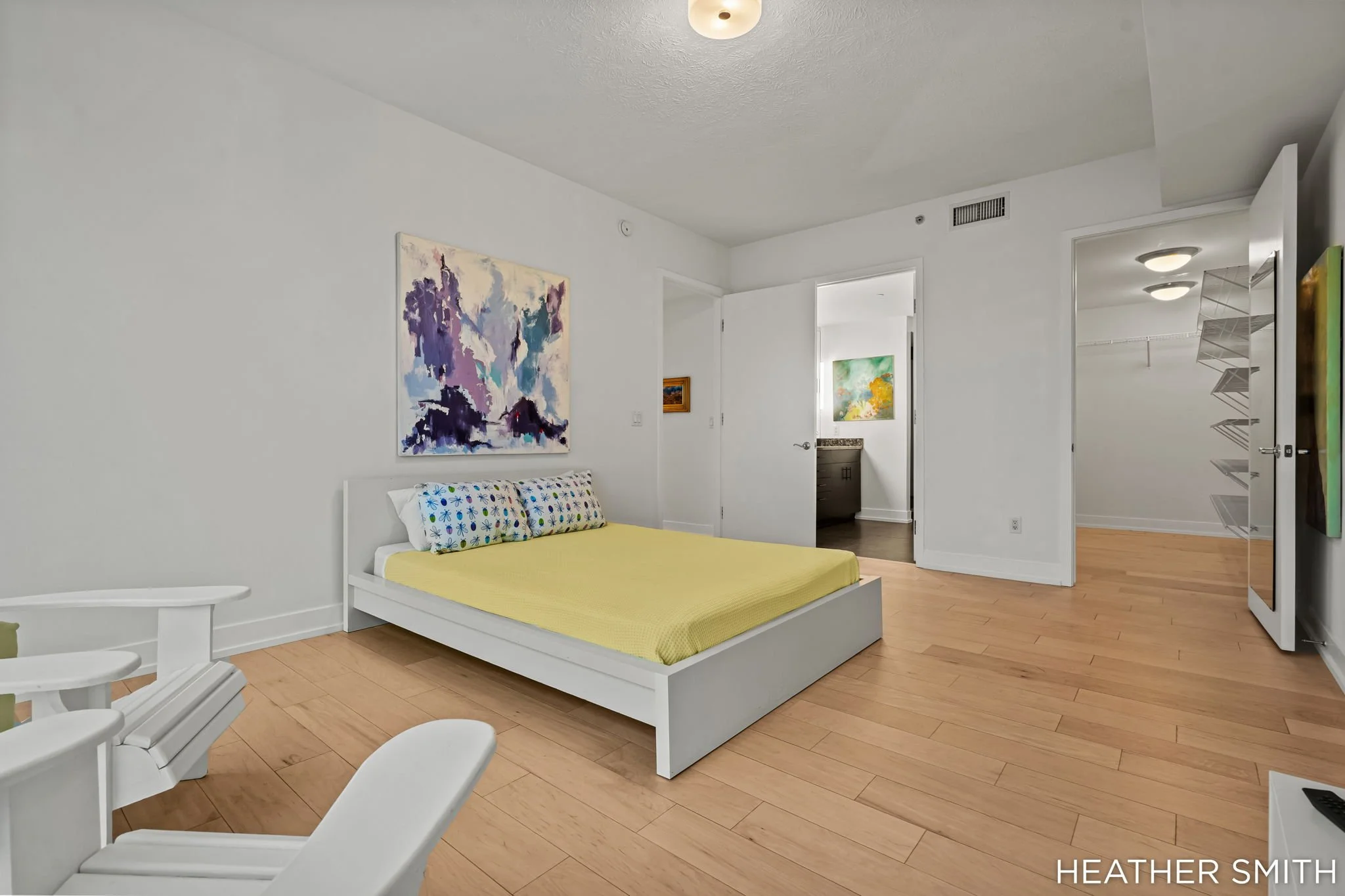A bedroom with light wood flooring, white walls, and a white bed with a yellow blanket and decorated pillows. There is a colorful abstract painting on the wall above the bed. The door to a closet and an open door to a bathroom are visible, with art o