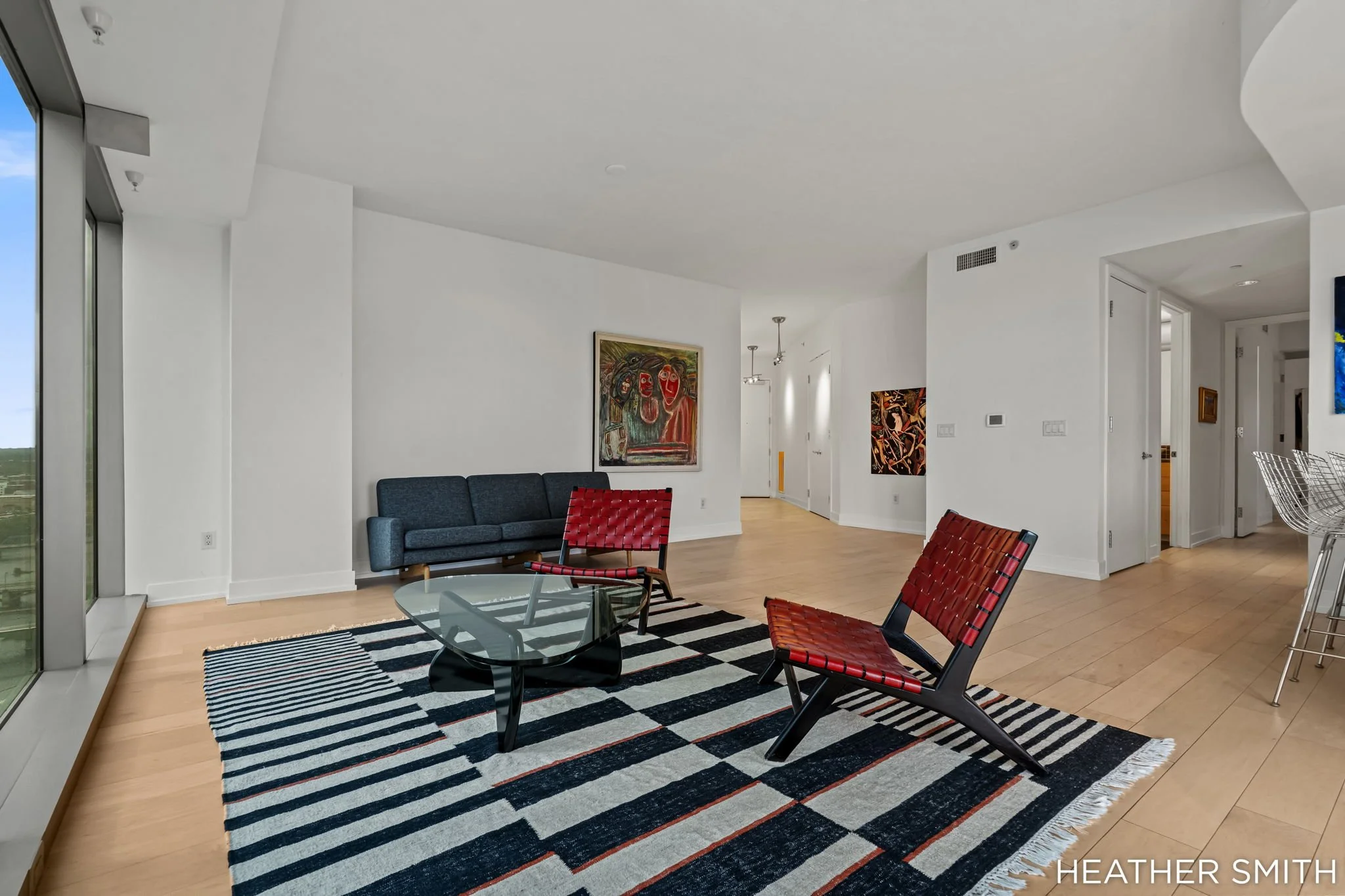 Modern living room with white walls, hardwood floors, a black and white striped rug, contemporary furniture including a blue sofa and red woven chairs, wall art, and large windows.