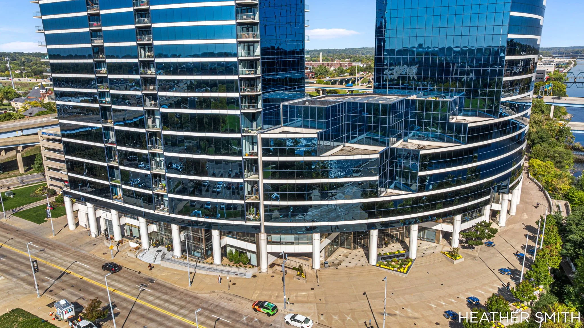 A modern, curved glass office building with multiple floors, balconies, and reflective windows. The building is situated in an urban area with roads, cars, greenery, and a river visible in the background.