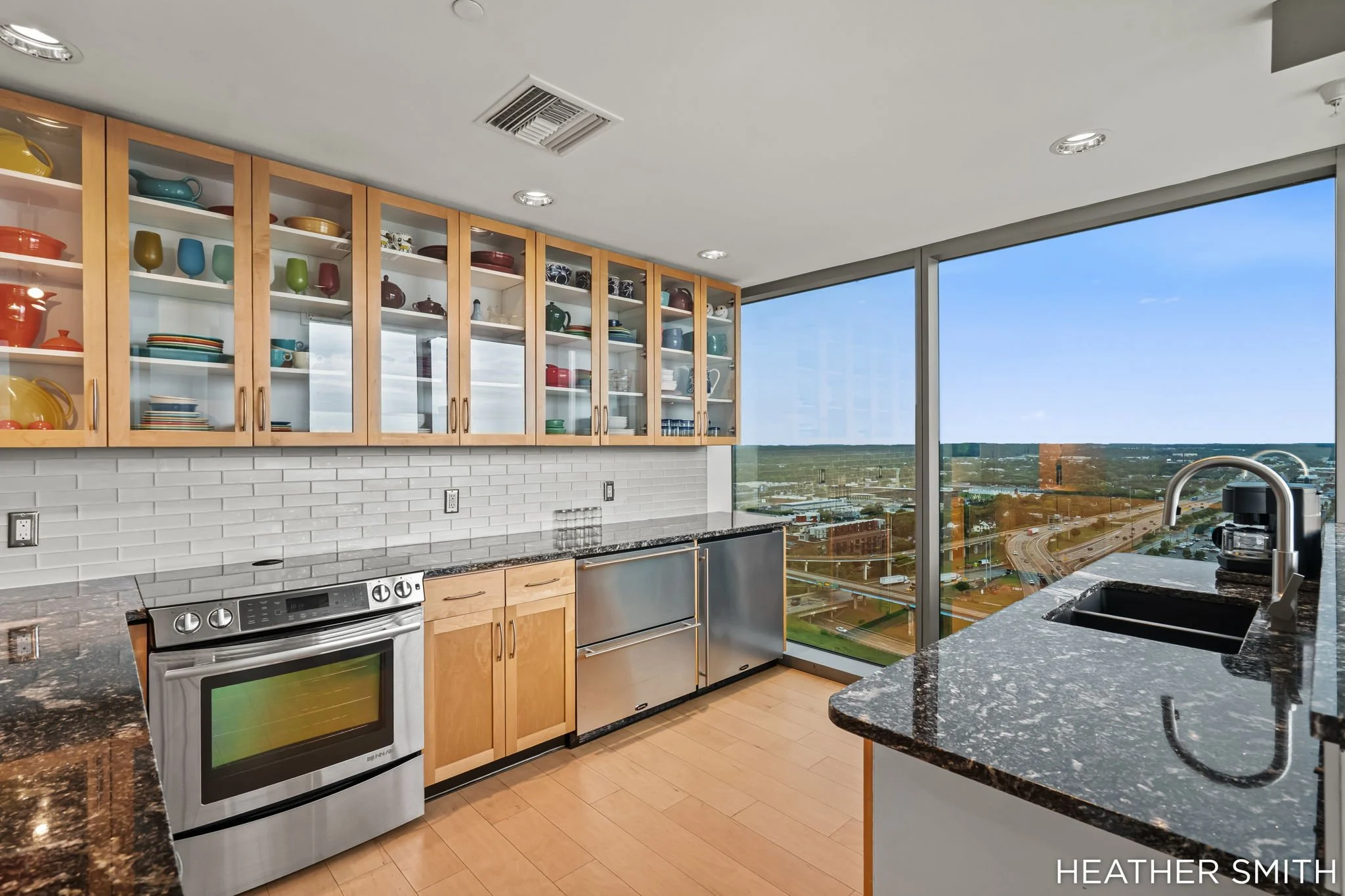 Modern kitchen with wooden cabinets and glass-front upper cabinets containing colorful dishware, large windows with a city view, granite countertops, a white subway tile backsplash, and stainless steel appliances.