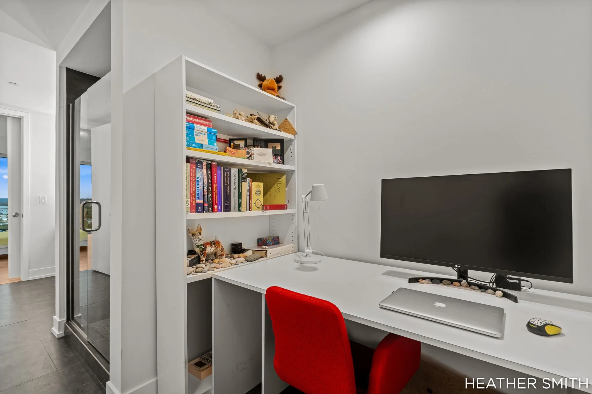 A modern home office with a white desk, a black computer monitor, a red chair, a white bookshelf filled with books and decorative items, and a small white lamp. There is a large window in the background with a view outside. The room has a minimalist 
