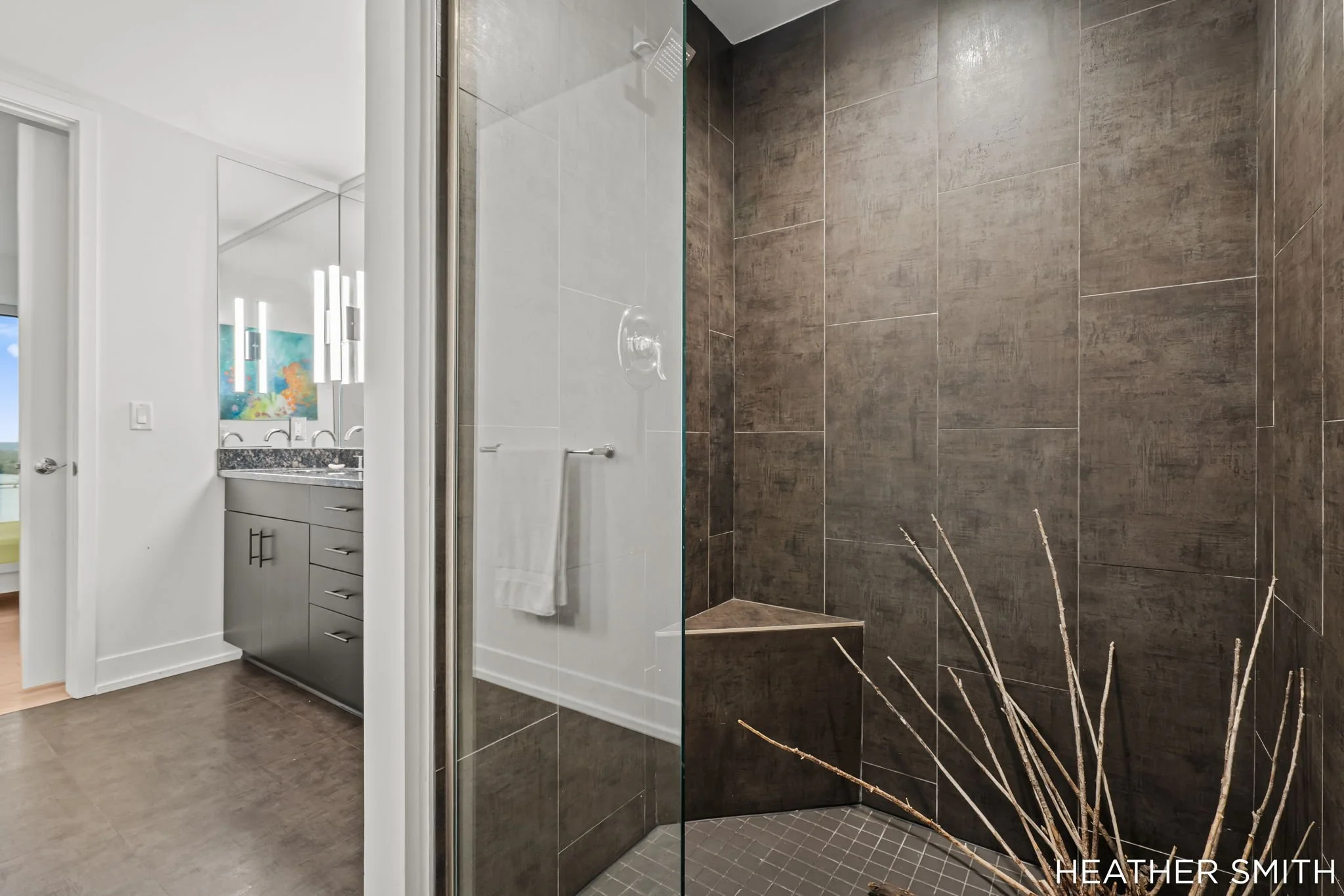 Modern bathroom with walk-in shower featuring dark brown tiles, a glass door, and a white towel; partial view of a vanity with a granite countertop and a colorful painting on the wall.