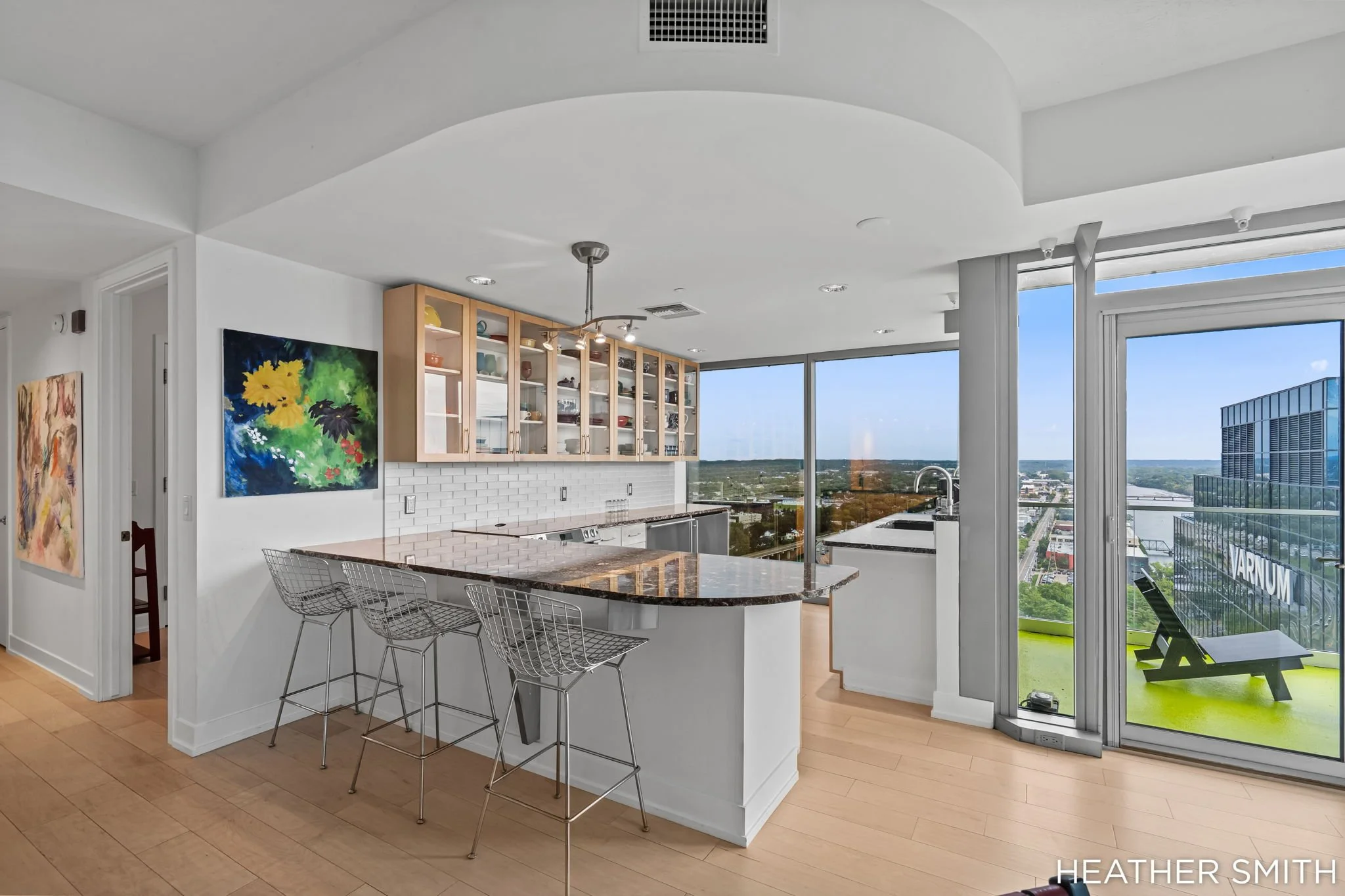 Modern kitchen with white walls, a curved marble countertop with bar stools, wooden upper cabinets with glass doors, large windows revealing a cityscape, and colorful artwork on the wall.
