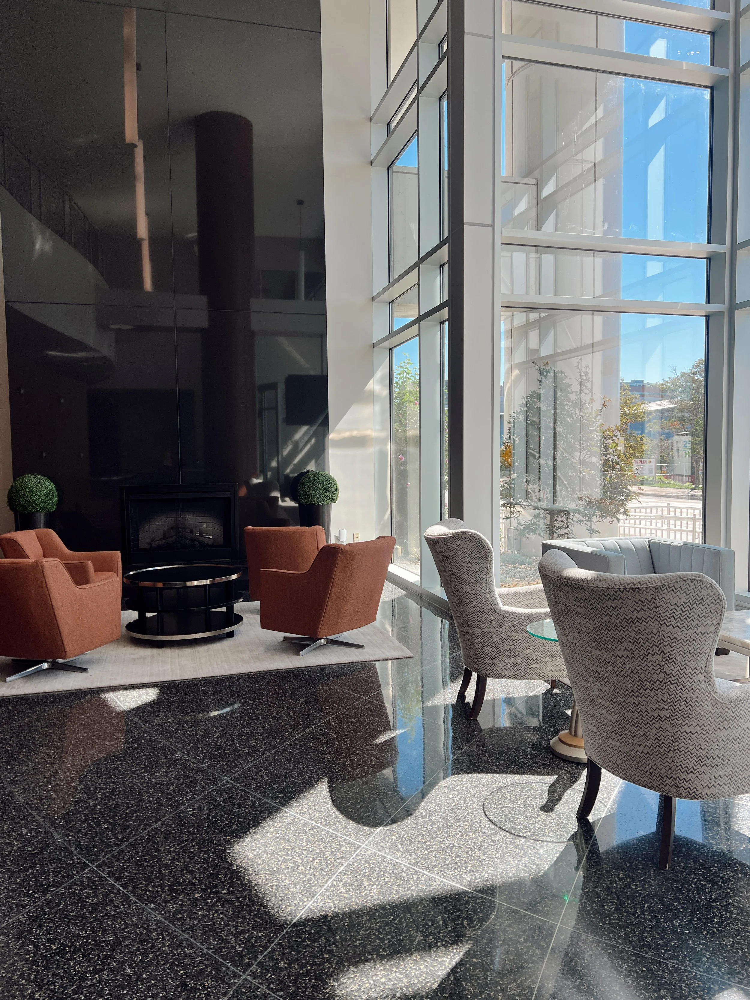 Interior of a modern building lobby with large floor-to-ceiling windows, sunlight casting shadows on the shiny black speckled tile floor, and seating including orange armchairs, patterned armchairs, and small glass tables.