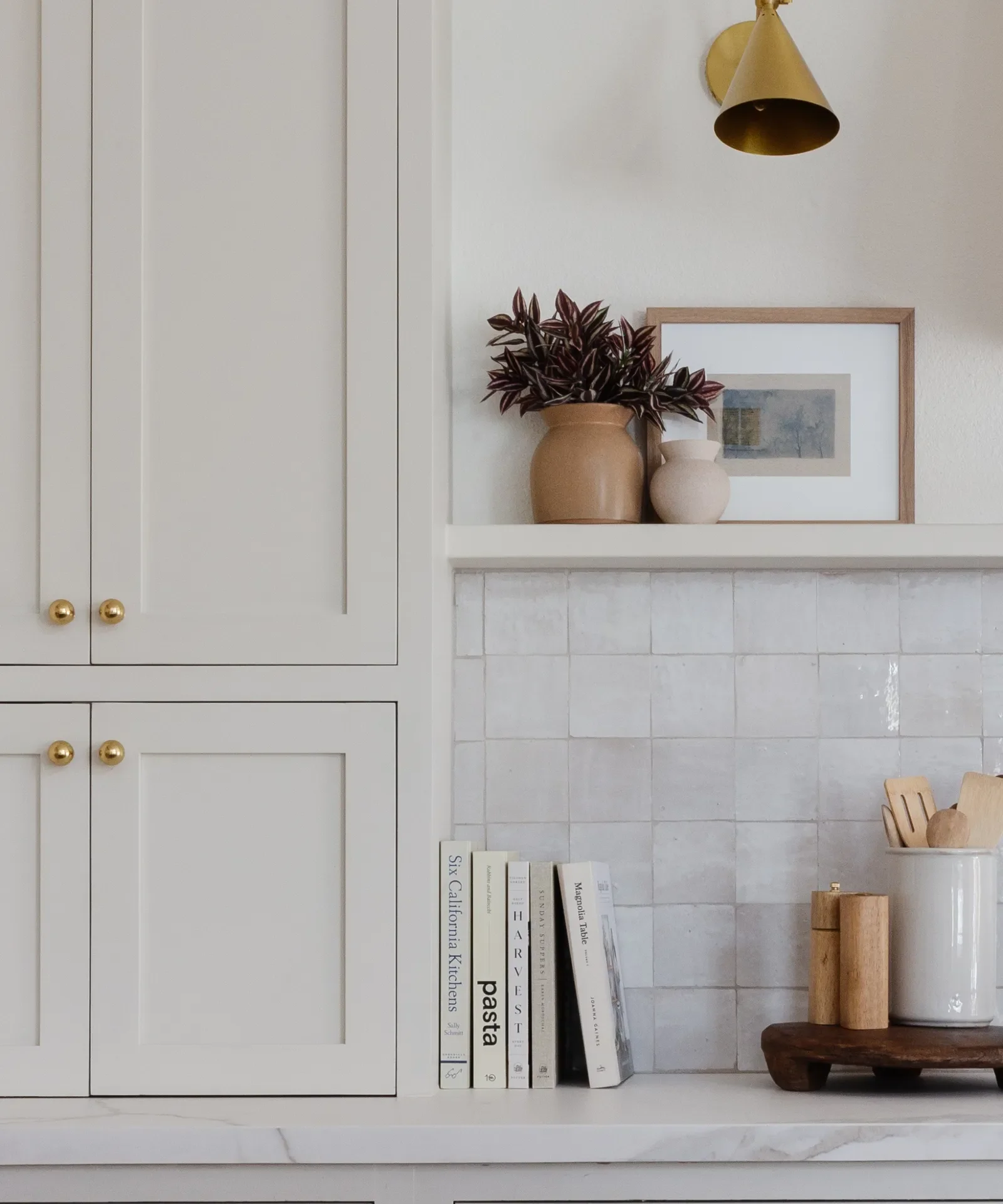 A kitchen wall with white cabinet doors, a shelf with two vases and framed artwork, and a countertop with books, a wooden cutting board, and a container with kitchen utensils.