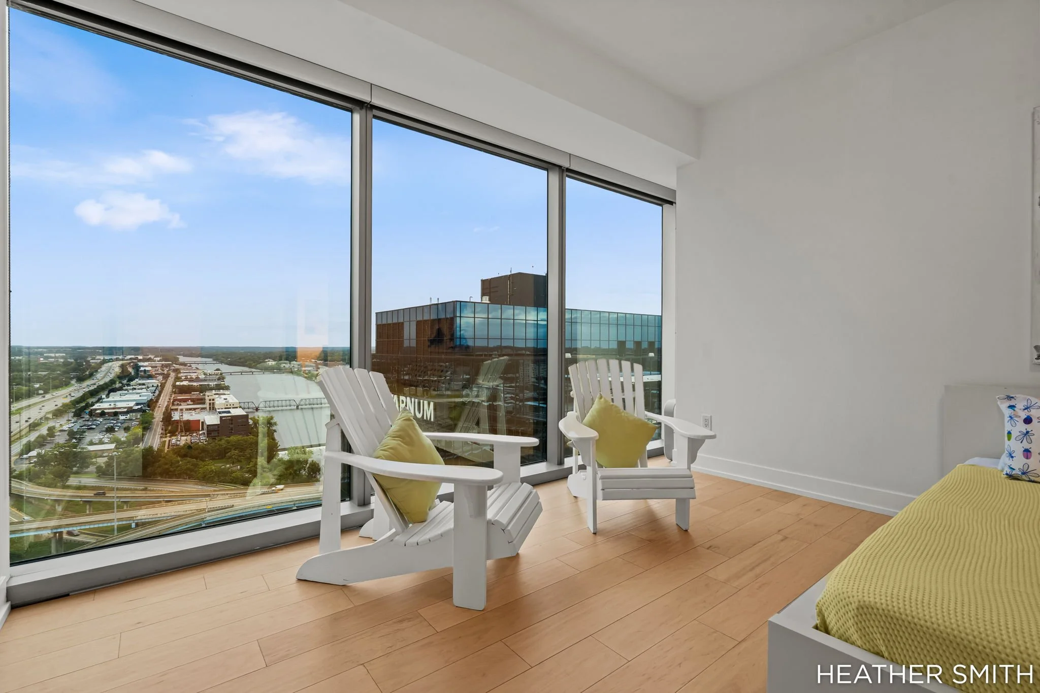 Interior of a modern apartment with large floor-to-ceiling windows showing a cityscape view, white chairs with yellow pillows, and a corner of a bed with a yellow bedspread.