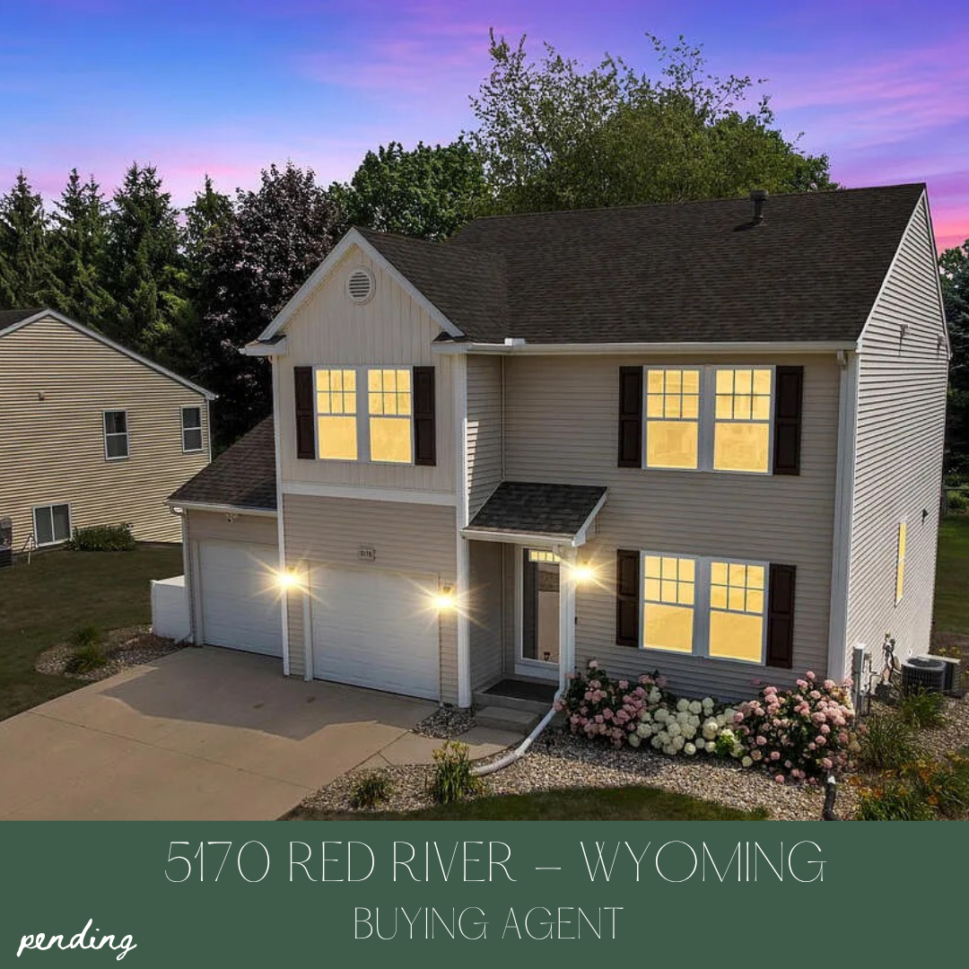 A two-story house with white siding and dark shutters, lit from inside, with a front yard flower bed, driveway, and neighboring house in Wyoming during sunset.