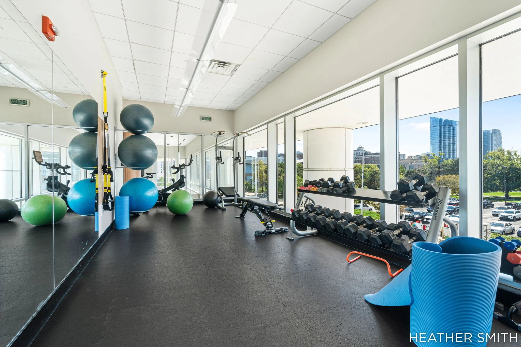 Fitness room with large windows, exercise balls, dumbbells, weight bench, and workout equipment, overlooking a cityscape with tall buildings.