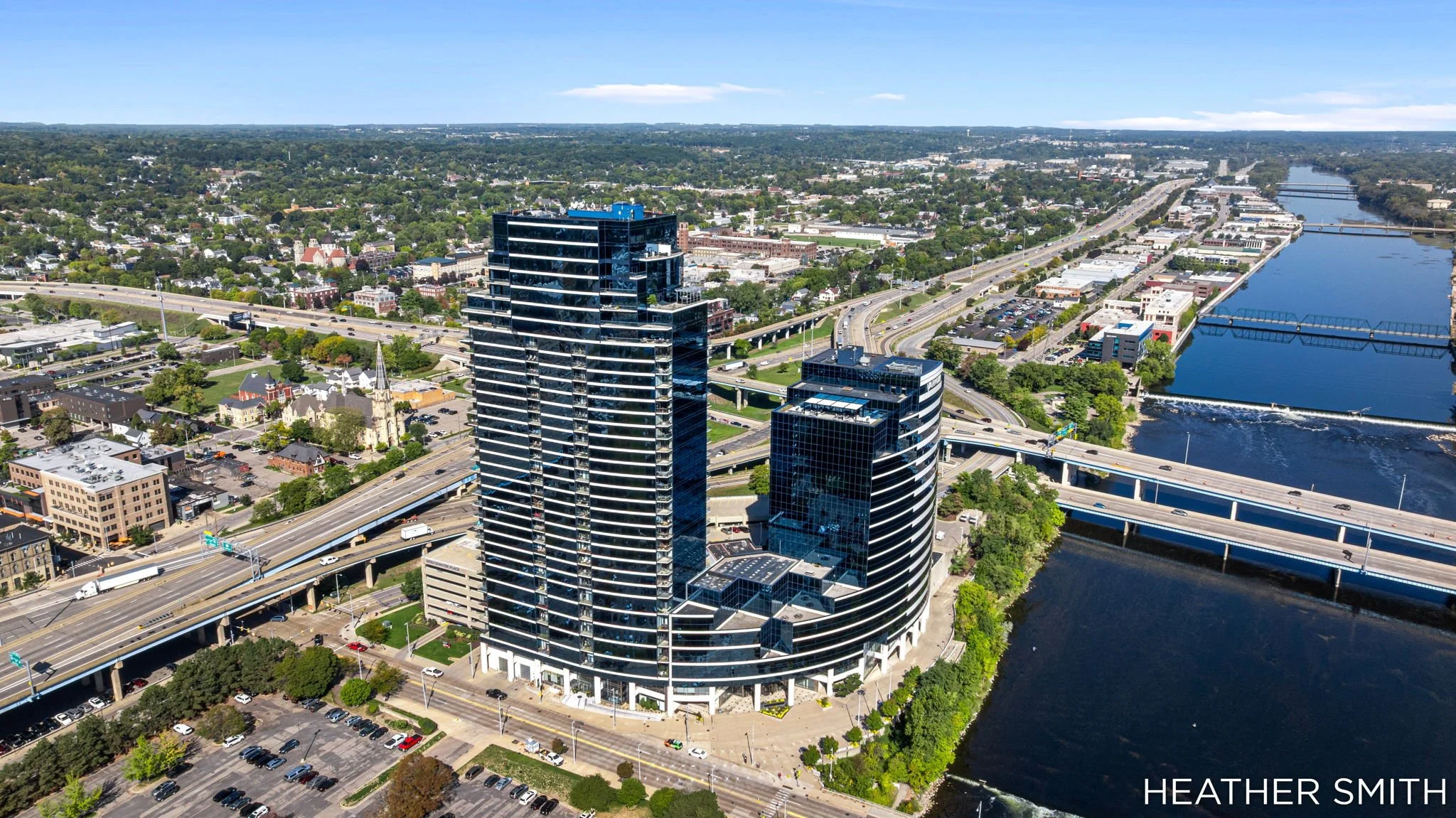 Aerial view of downtown Nashville, Tennessee, featuring modern high-rise buildings, highways, a river, and surrounding cityscape under a clear blue sky.
