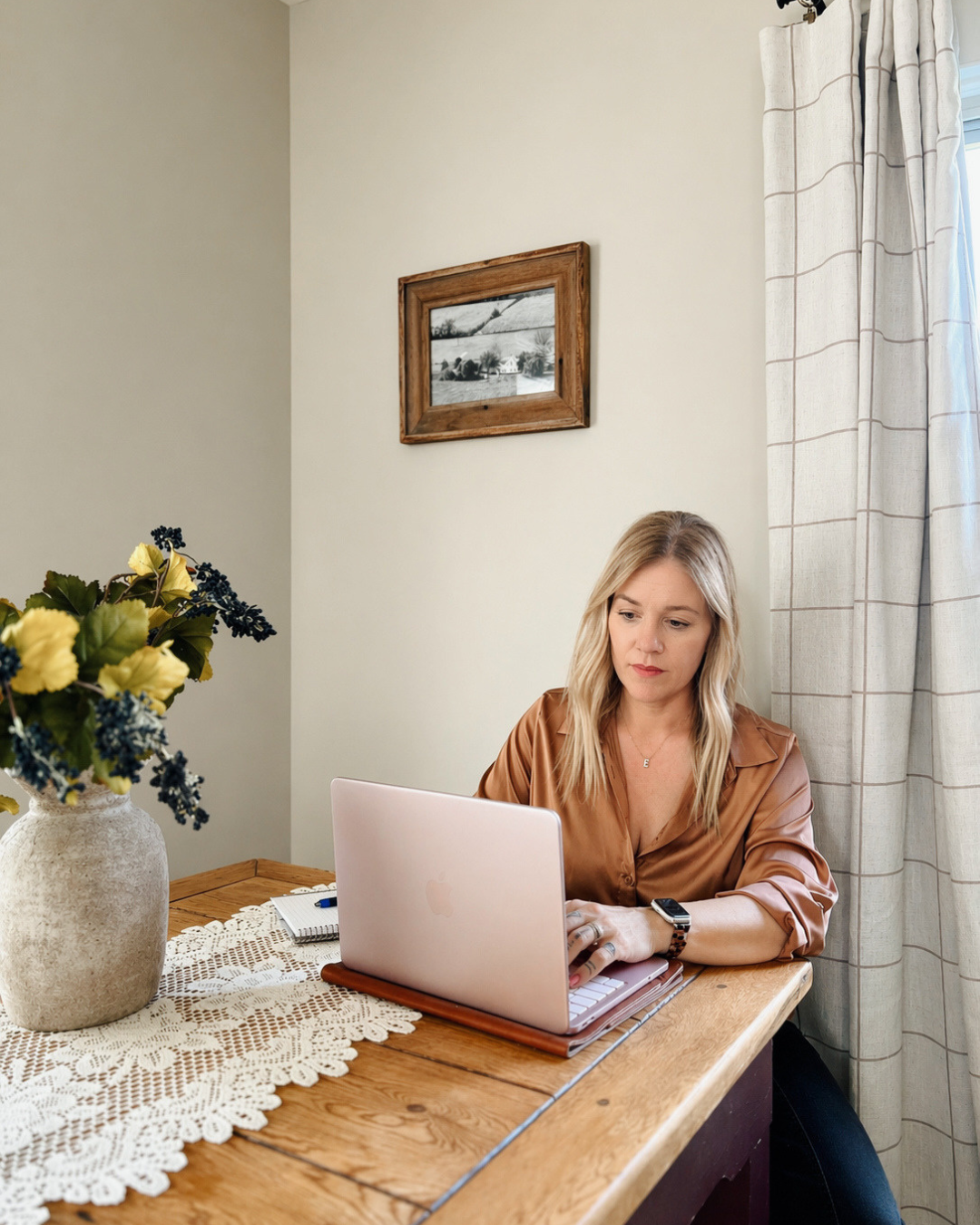 A woman with blonde hair working on a silver laptop at a wooden dining table, with a vase of flowers, a notebook, and a pen nearby, in a room with beige walls, a framed picture, and curtains.
