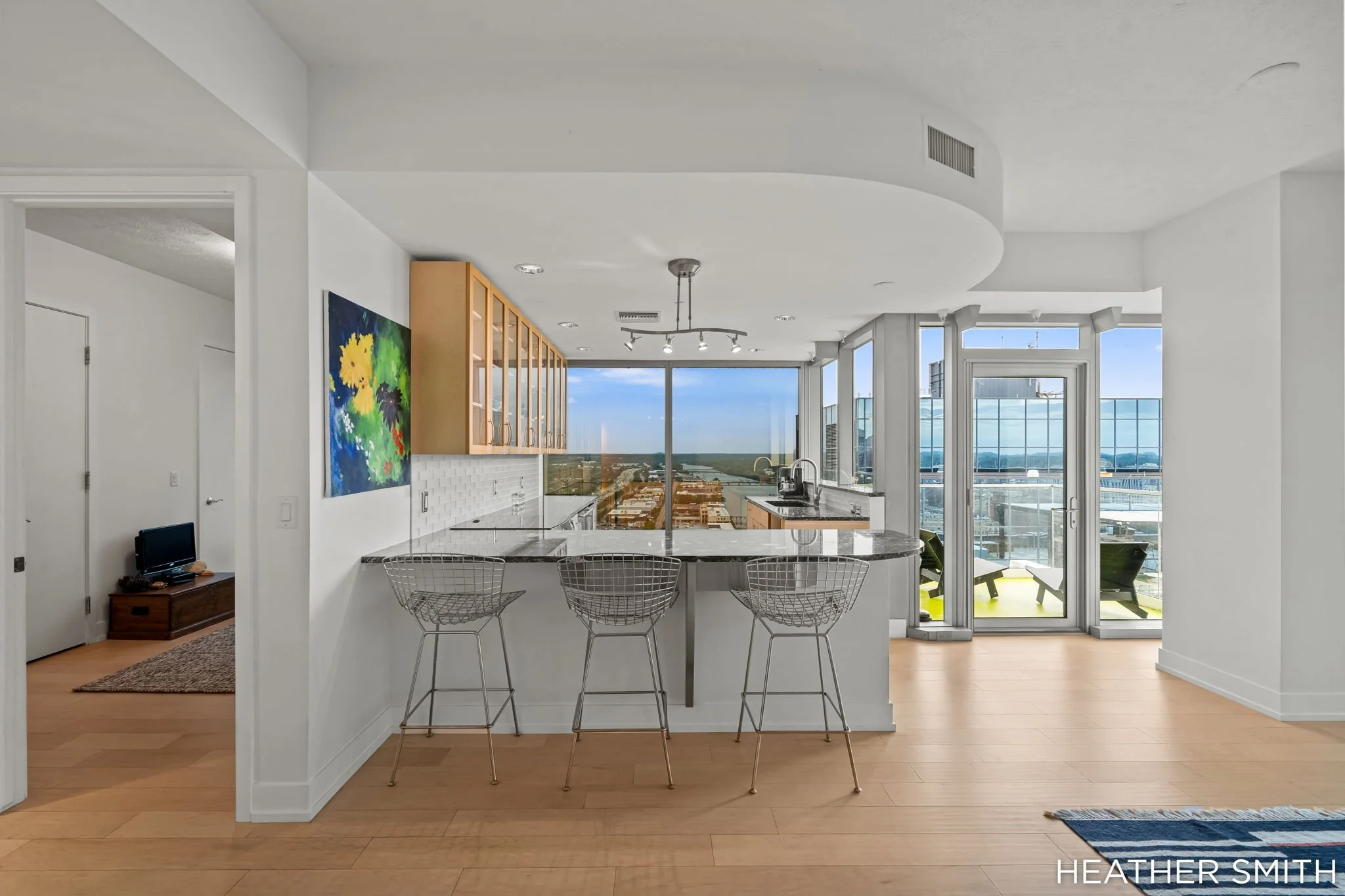 Modern kitchen and living space with panoramic window view, island with bar stools, and artwork on the wall.