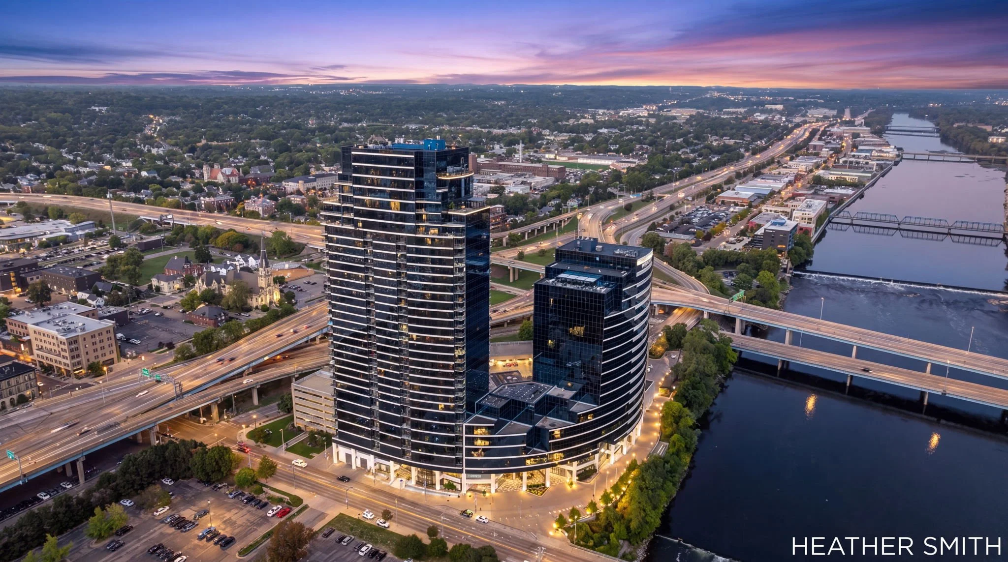 Aerial view of a cityscape during sunset, featuring tall modern glass buildings, highways, a river, and green areas.