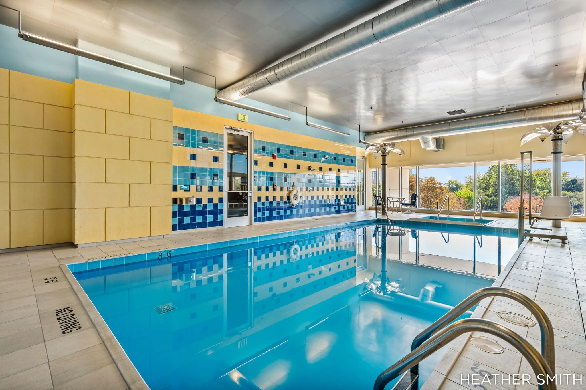 Indoor swimming pool with blue and yellow tiled walls, large windows showing trees outside, and swimming area with seating and pool equipment.