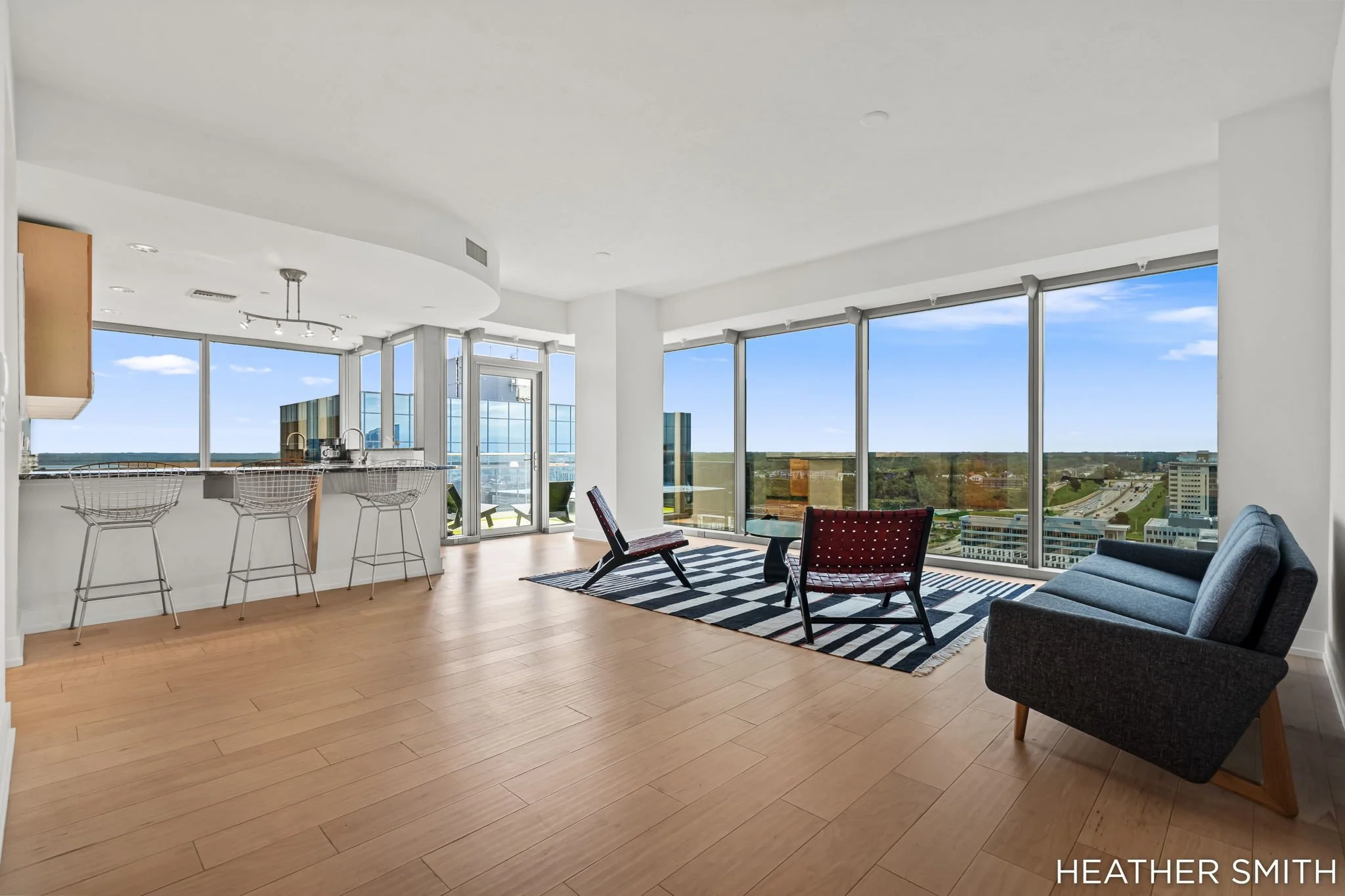 A modern high-rise apartment living room with large floor-to-ceiling windows showing a city view. The room has wooden flooring, a gray sofa, two red chairs, a black and white striped rug, a small side table, and a kitchen area with bar stools and a c