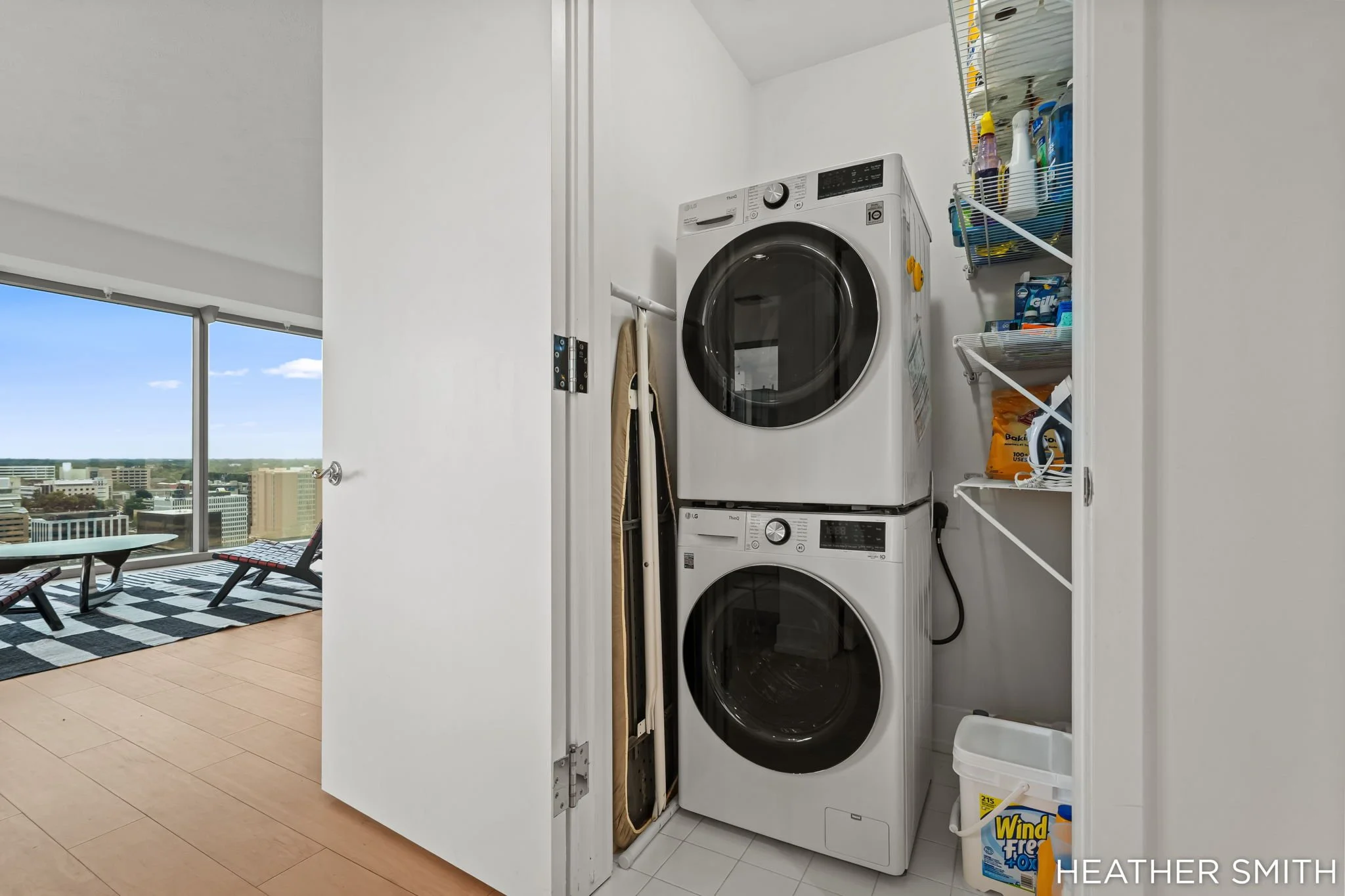 A laundry closet with a stacked washer and dryer, cleaning supplies on a shelf, and a window nearby showing a cityscape view.