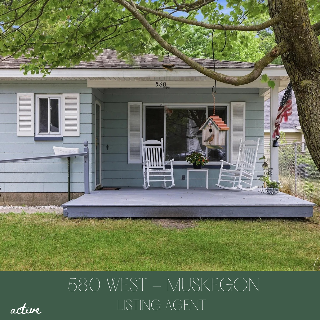 Front porch of a house with light blue siding, white window shutters, a small deck with white rocking chairs, a hanging birdhouse, an American flag, and a tree with green leaves.