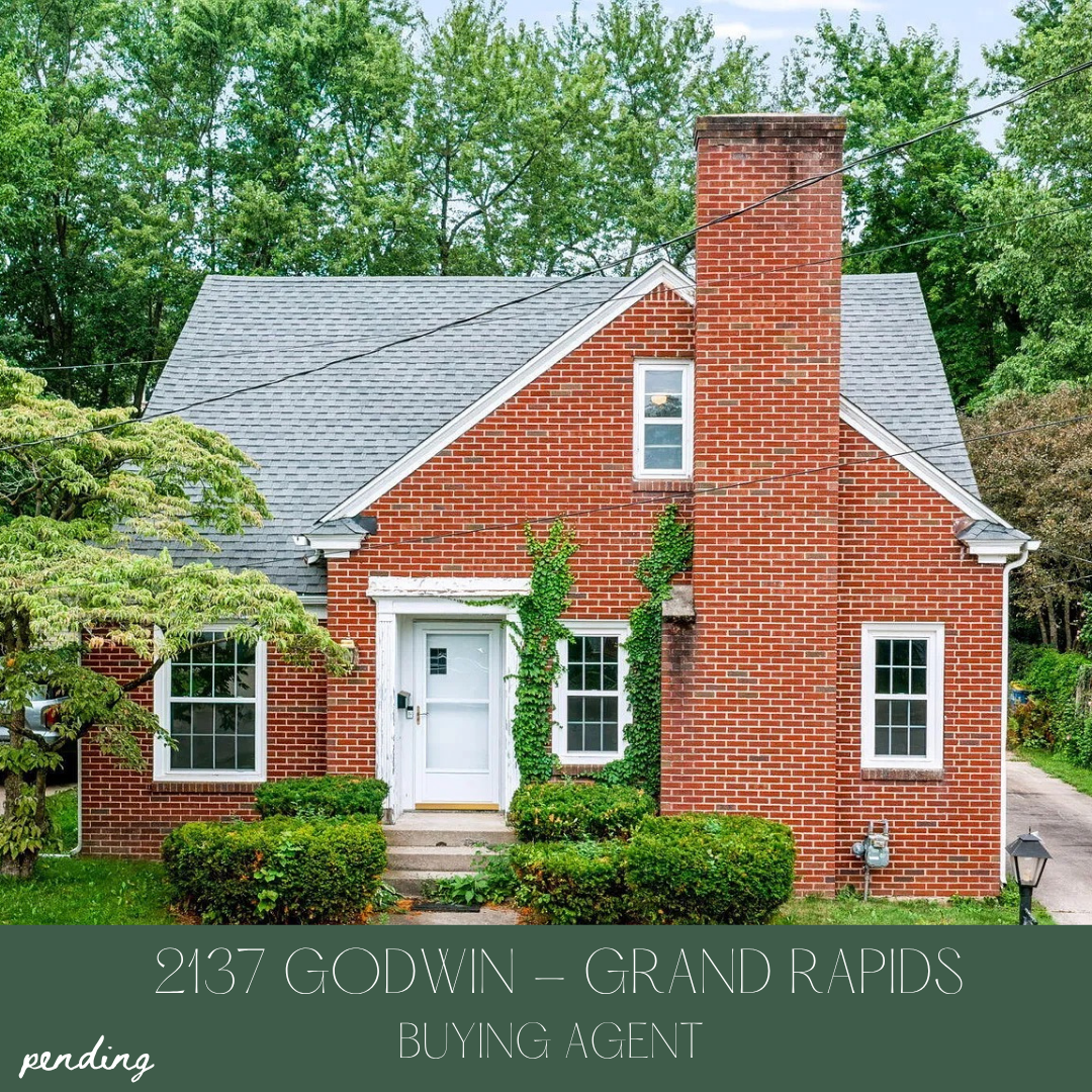 A brick house with a gray shingle roof, white front door, and ivy growing on the brick wall, surrounded by greenery.