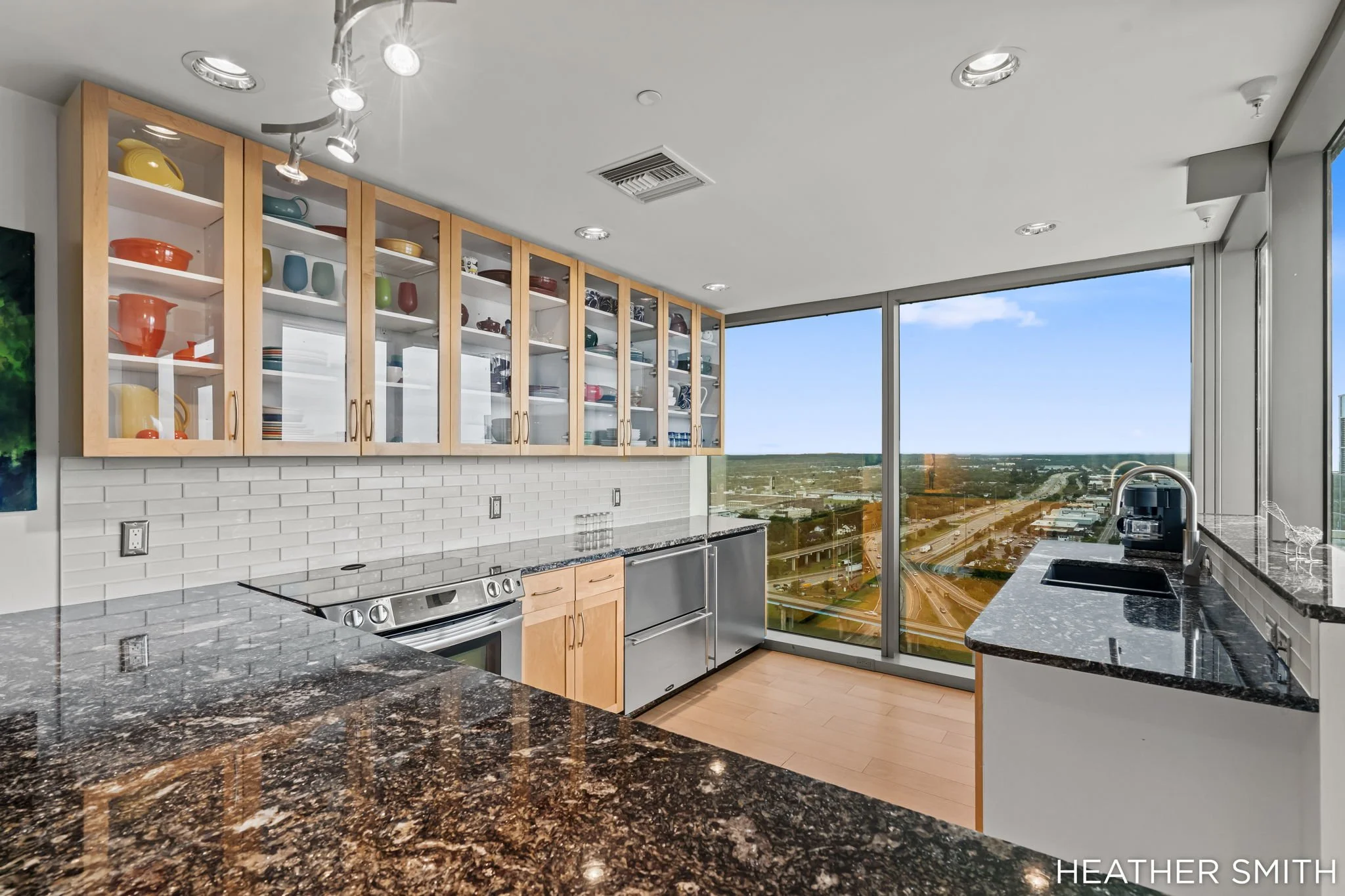 Modern kitchen with open cabinets displaying colorful dishes, granite countertops, stainless steel appliances, and large windows showcasing a city view.