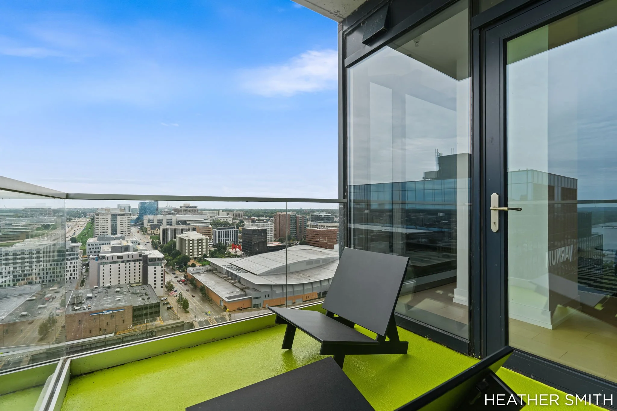 View from a high-rise balcony showing a cityscape with various buildings, streets, and a clear blue sky. The balcony has a glass railing and a black-framed glass door, with a black chair facing the view. The wall near the door is painted green, and t