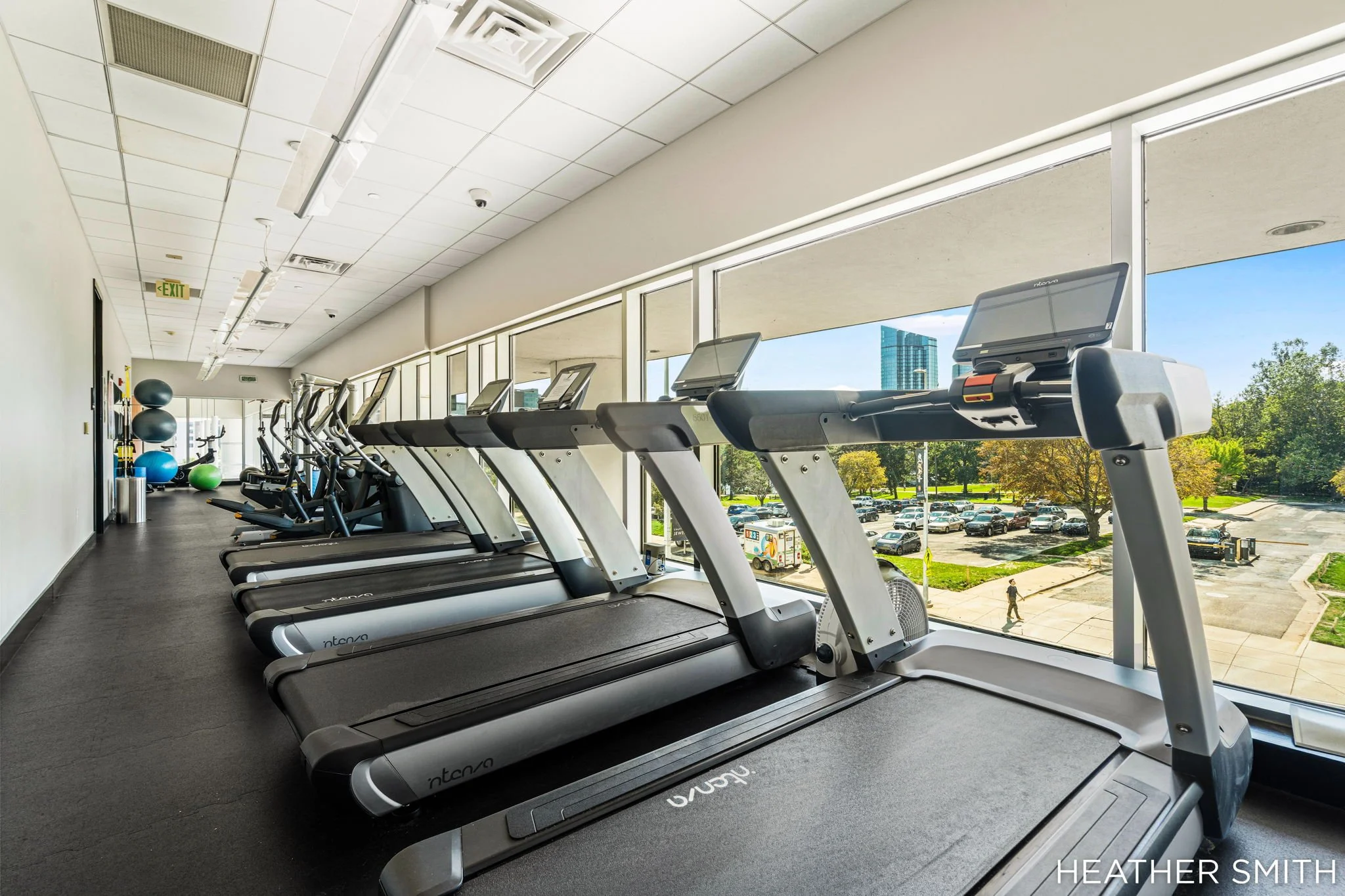 Indoor gym with several treadmills facing large windows overlooking a parking lot with cars and trees outside. Exercise balls are stacked along the wall to the left. The ceiling has air vents and lighting fixtures. Bright daylight outside.