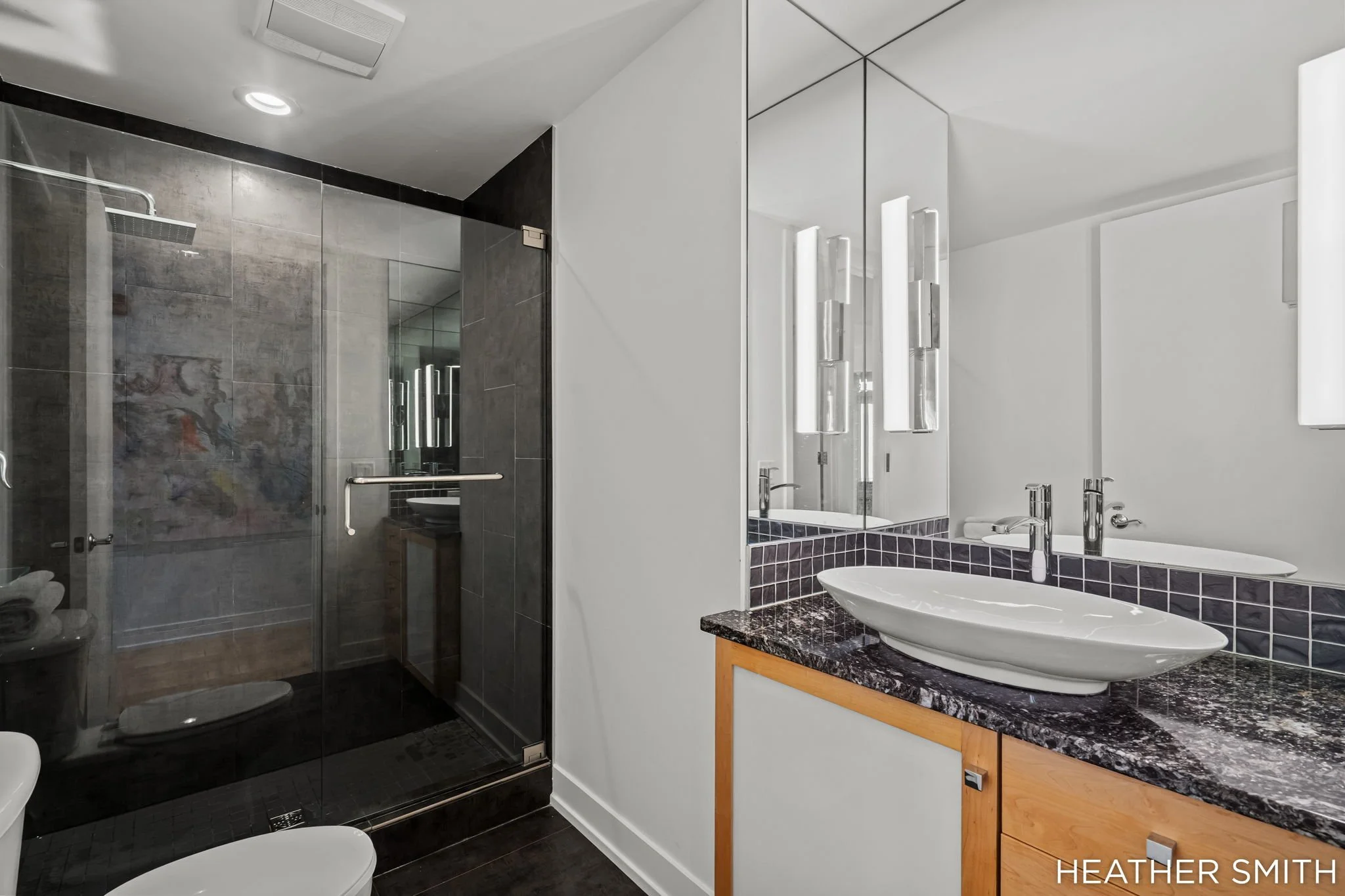 Modern bathroom with a glass-enclosed shower, double sink vanity with black granite countertop, and mirror with vertical lighting fixtures, featuring a white wall and black tiles.