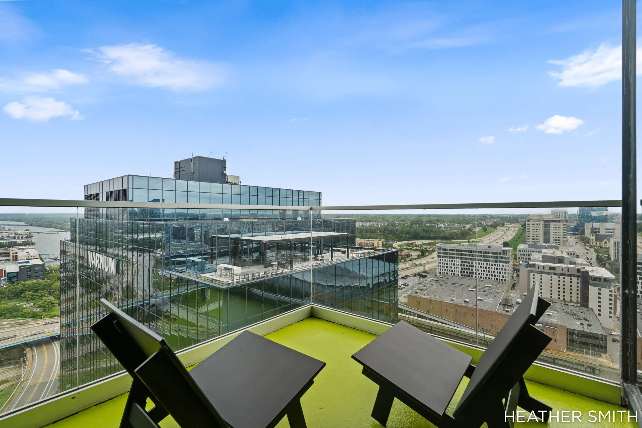 A rooftop balcony with three black tables and black chairs. The balcony has a glass railing, overlooking a cityscape with tall buildings, roads, and a river in the background under a bright blue sky with scattered clouds.