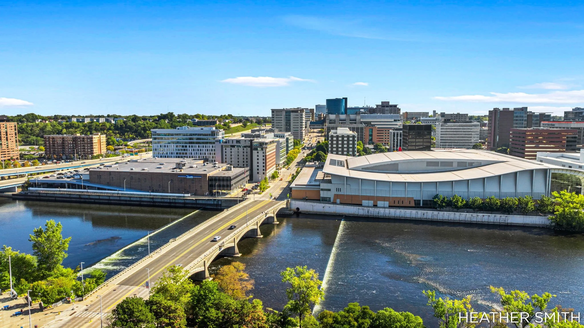 A cityscape with modern office buildings, a river with a bridge, and green trees on a sunny day.