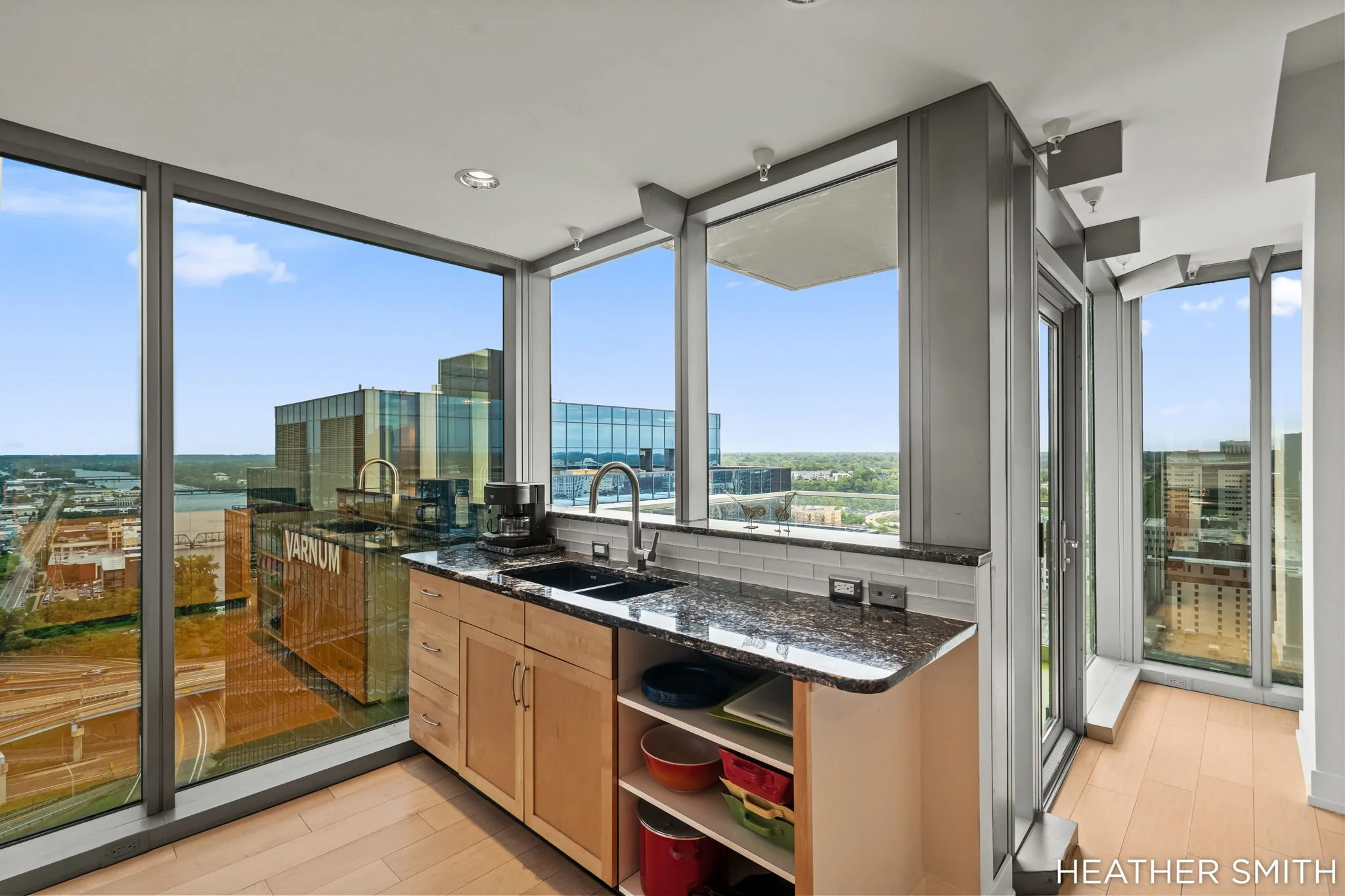Modern kitchen with large floor-to-ceiling windows offering city skyline view, light wood cabinets, black granite countertops, and a stainless steel sink.