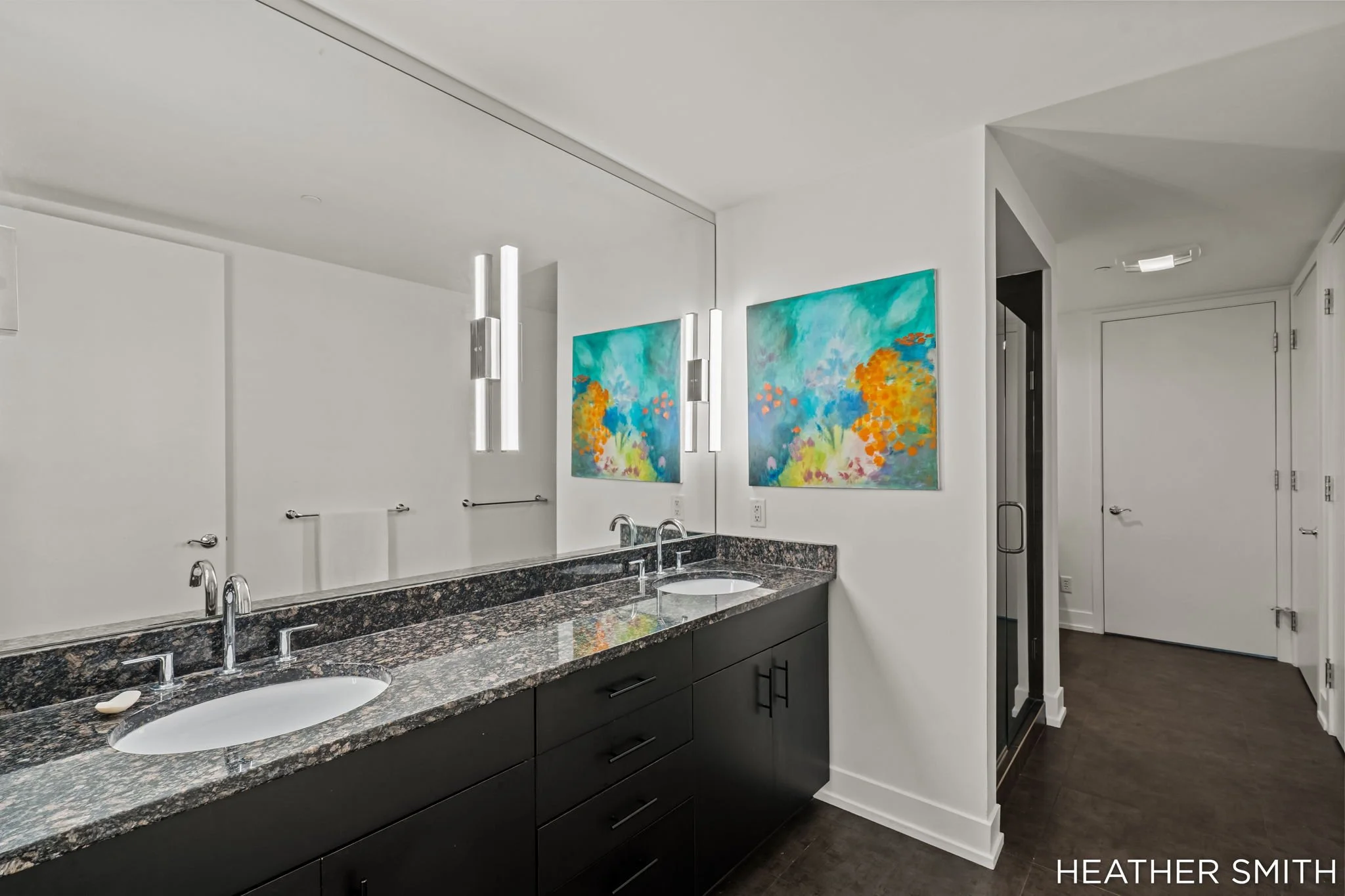 Modern bathroom with white walls, black cabinetry with silver handles, a granite countertop, two undermount sinks, and colorful abstract paintings on the wall.