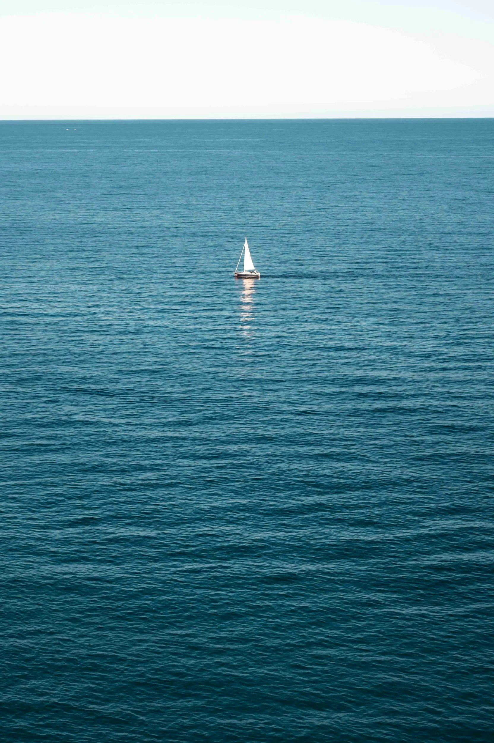 A sailboat on calm blue ocean waters with a light-colored sky in the background.