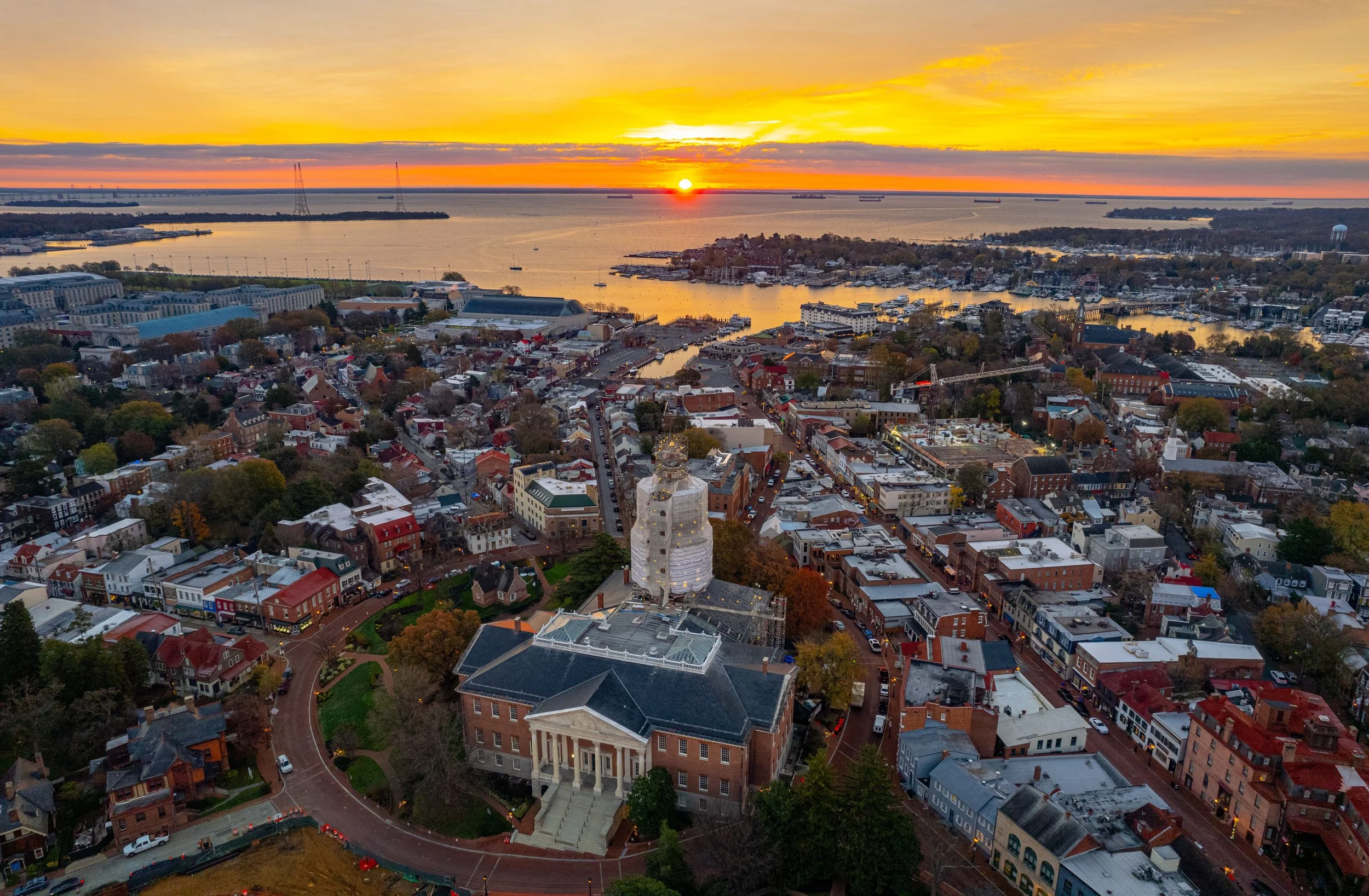 Aerial view of a city at sunset with a prominent historic building featuring a clock tower in the foreground, a river with boats, and a colorful sky with the setting sun.