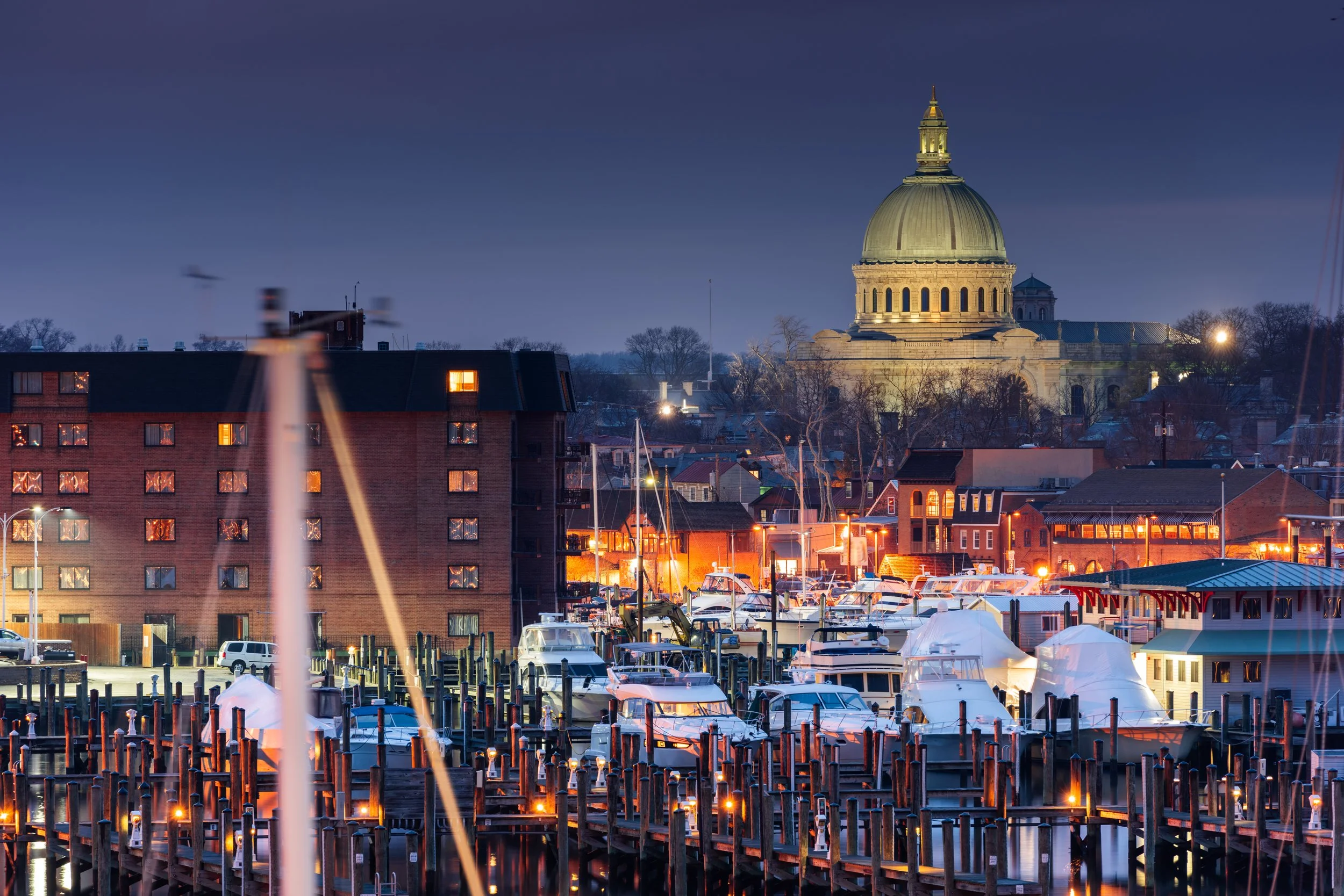 Night view of a harbor with docked boats and a cityscape, including a prominent historic building with a large dome, illuminated by streetlights.