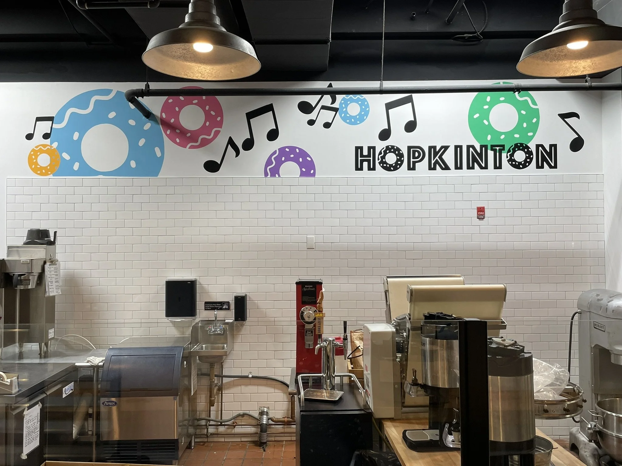 Interior of a cafe or bakery with a white tiled wall, colorful donut and musical note wall art, and the word 'HOPKINTON' displayed.