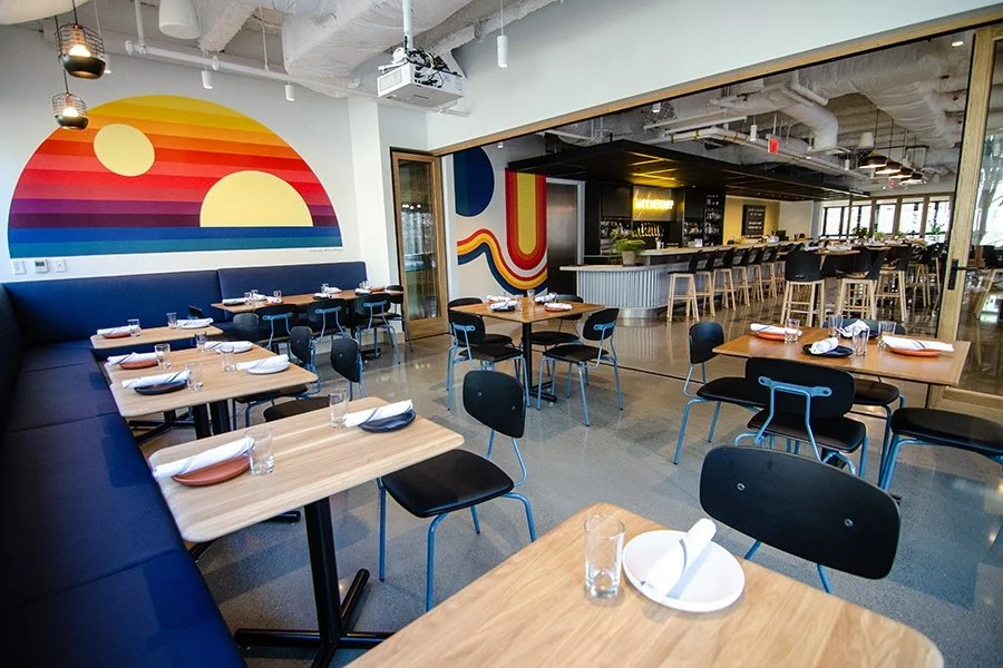 Modern restaurant interior with wooden tables, black chairs, and a colorful sunset mural on the wall.