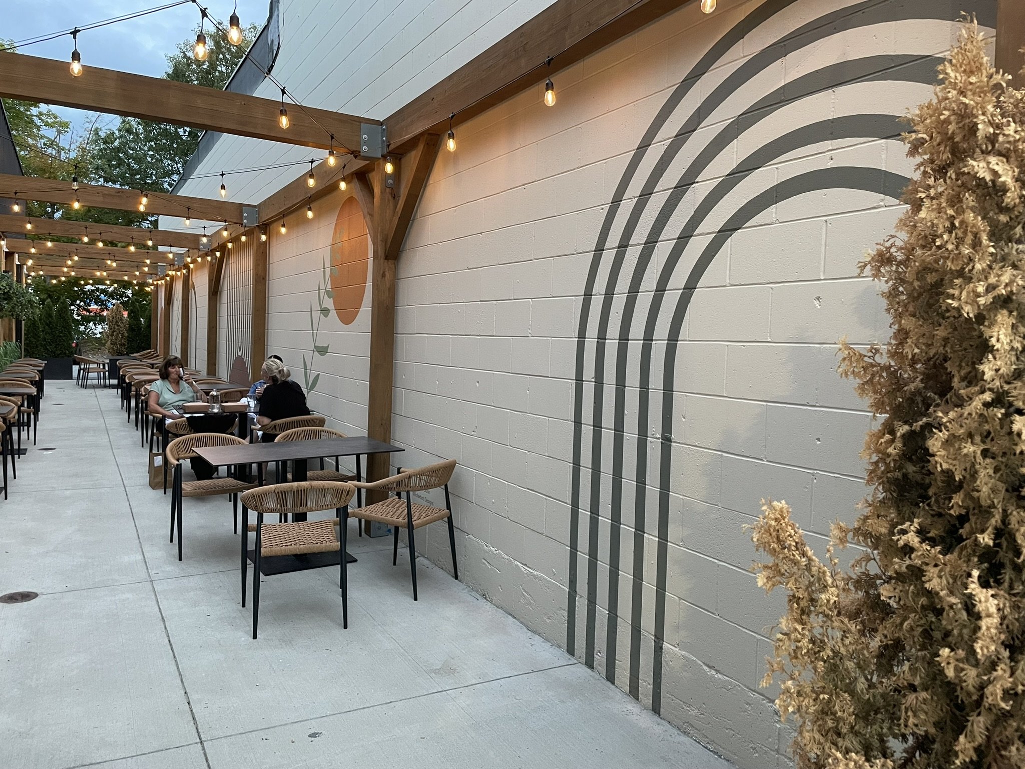 Outdoor patio with wooden beams, string lights, tables and chairs, two women talking, mural of a rainbow on the wall, plants, and a sidewalk.