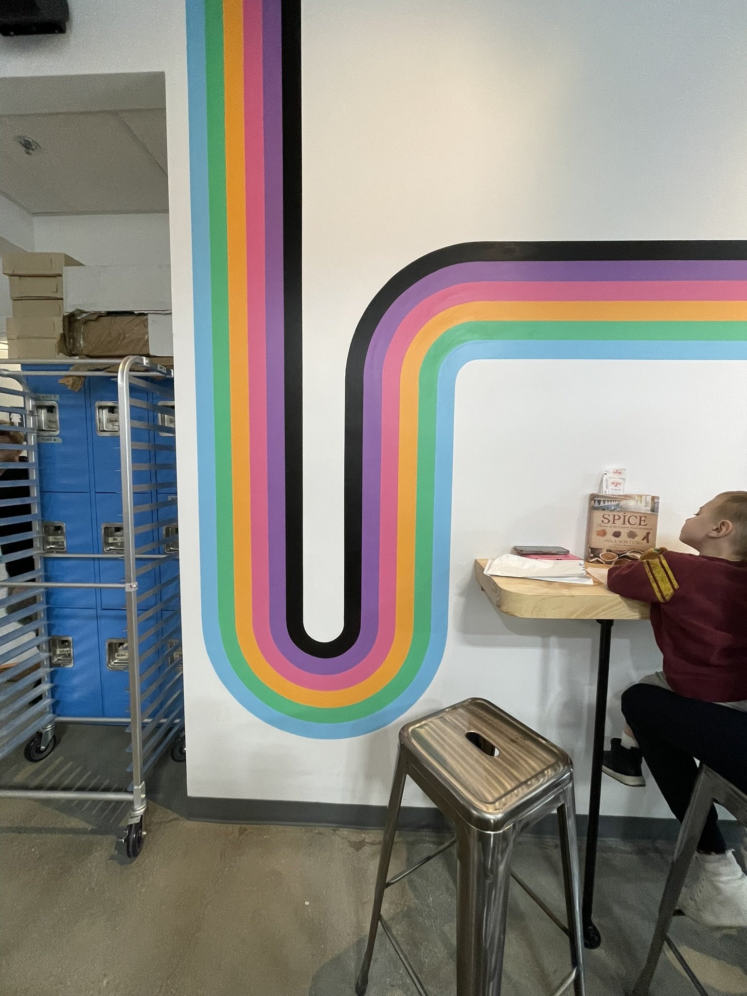 Colorful rainbow stripe mural on a wall inside a cafe, with a child sitting at a wooden table.