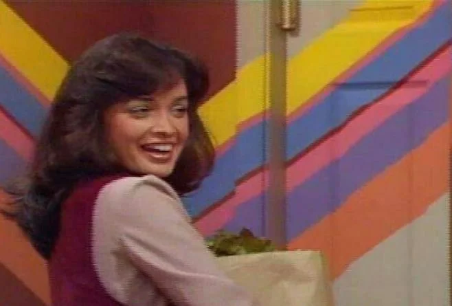 A woman with dark, shoulder-length hair smiling, holding a bag of groceries, in front of a colorful, striped wall.