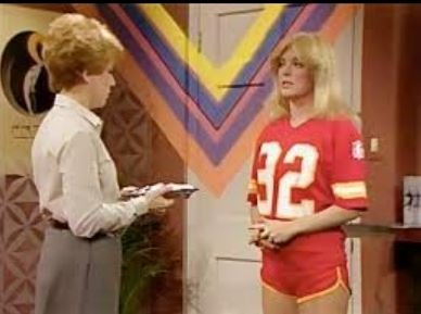 Two women in conversation indoors, one in a white shirt and gray pants, the other in a red sports jersey with the number 52.