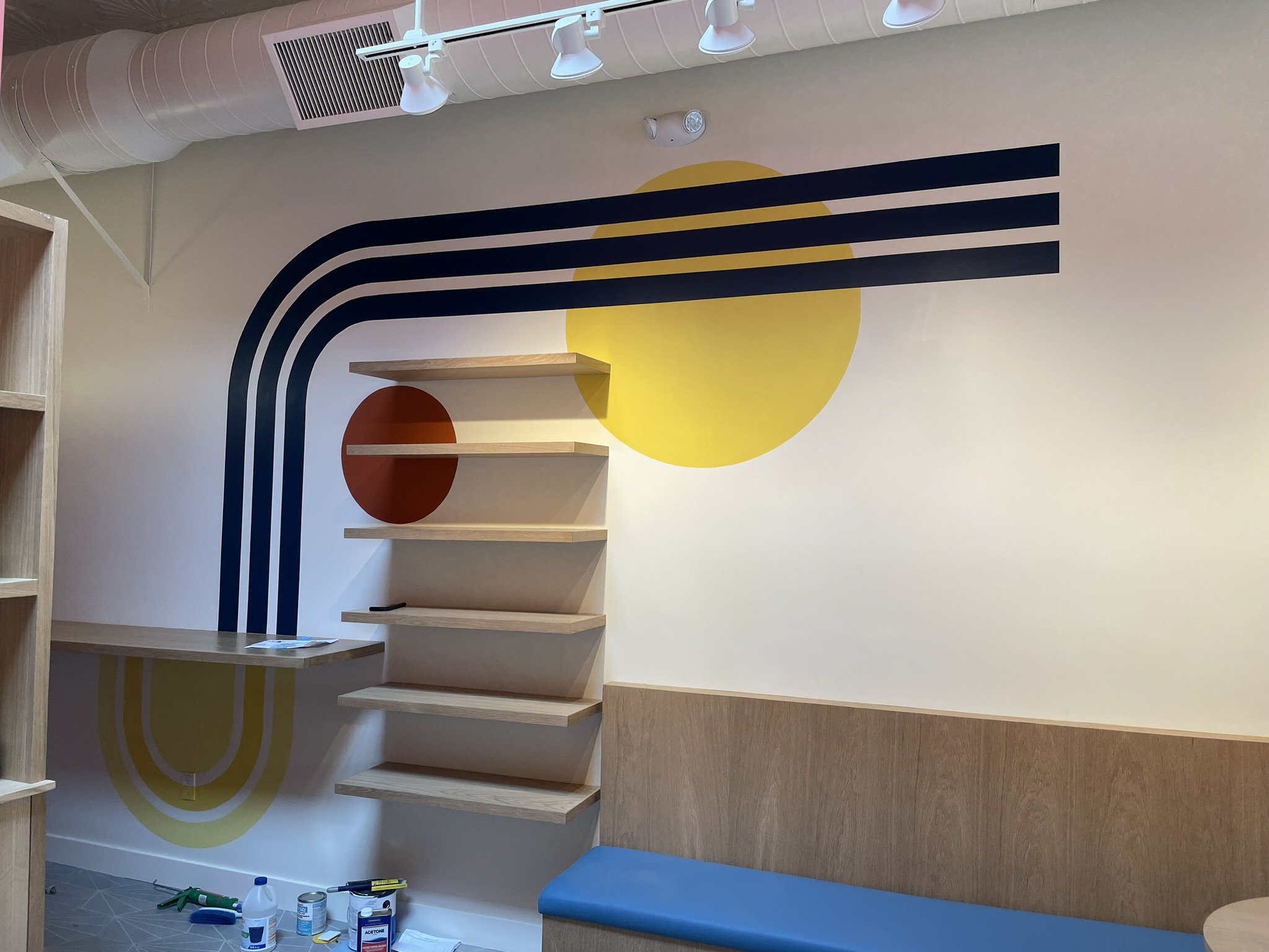 A room with colorful abstract wall art featuring black, yellow, orange, and blue shapes and lines. Wood shelving and a blue padded bench beneath the artwork. Some tools and supplies on the floor.