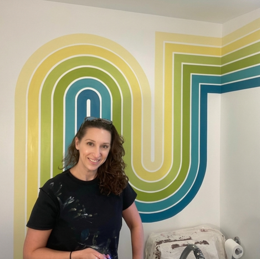 A woman with brown curly hair wearing a black shirt with paint splatters standing in front of a colorful retro-striped wall mural.