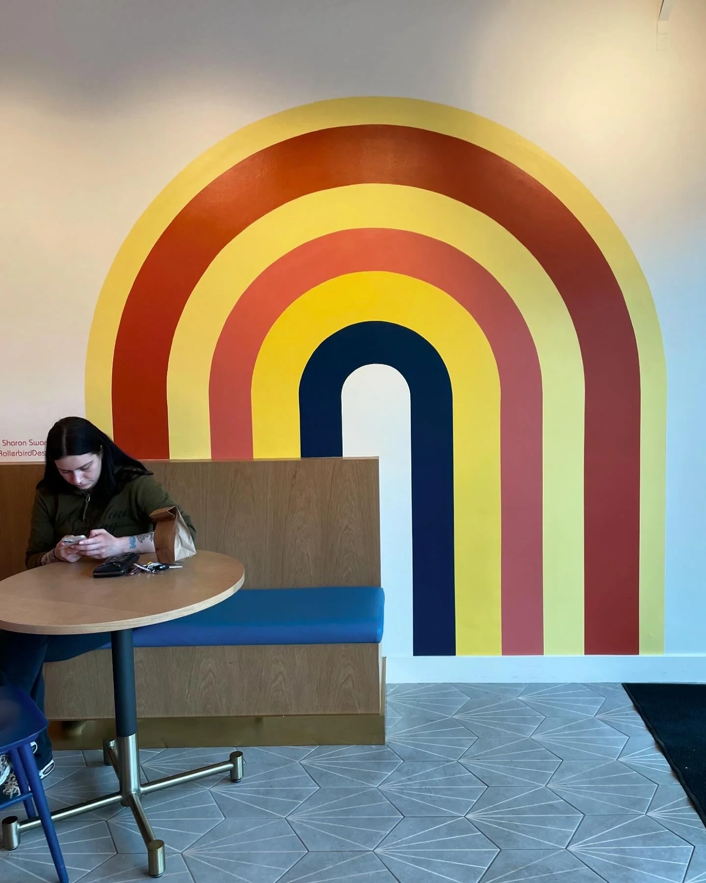 A woman sitting at a table in a cafe, looking at her phone, with a colorful rainbow mural on the wall behind her.