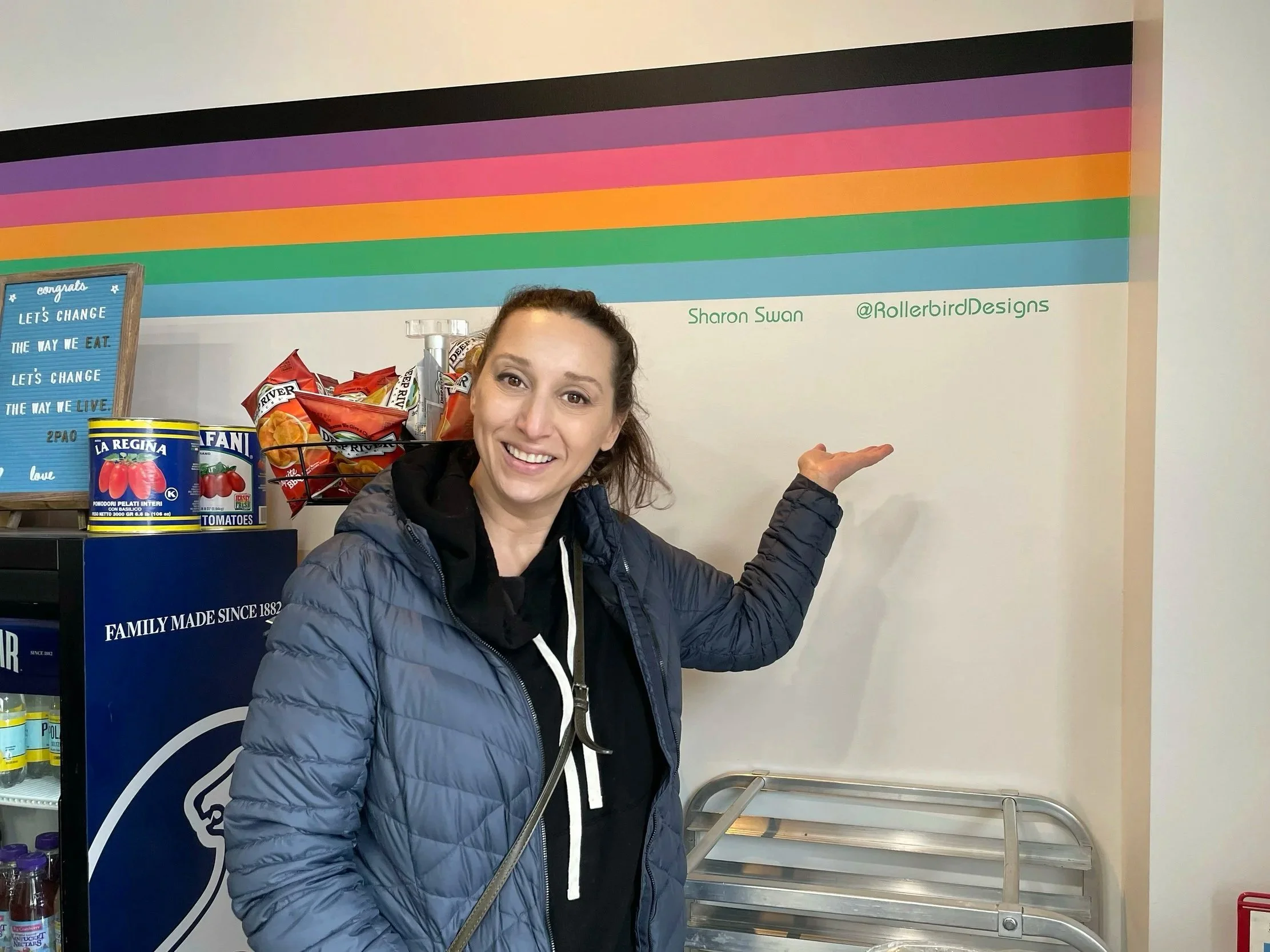 A woman in a blue jacket standing in front of a colorful rainbow wall, pointing to her right. The wall has the name 'Sharon Swann' and the Instagram handle '@RollerbirdDesigns' written on it.