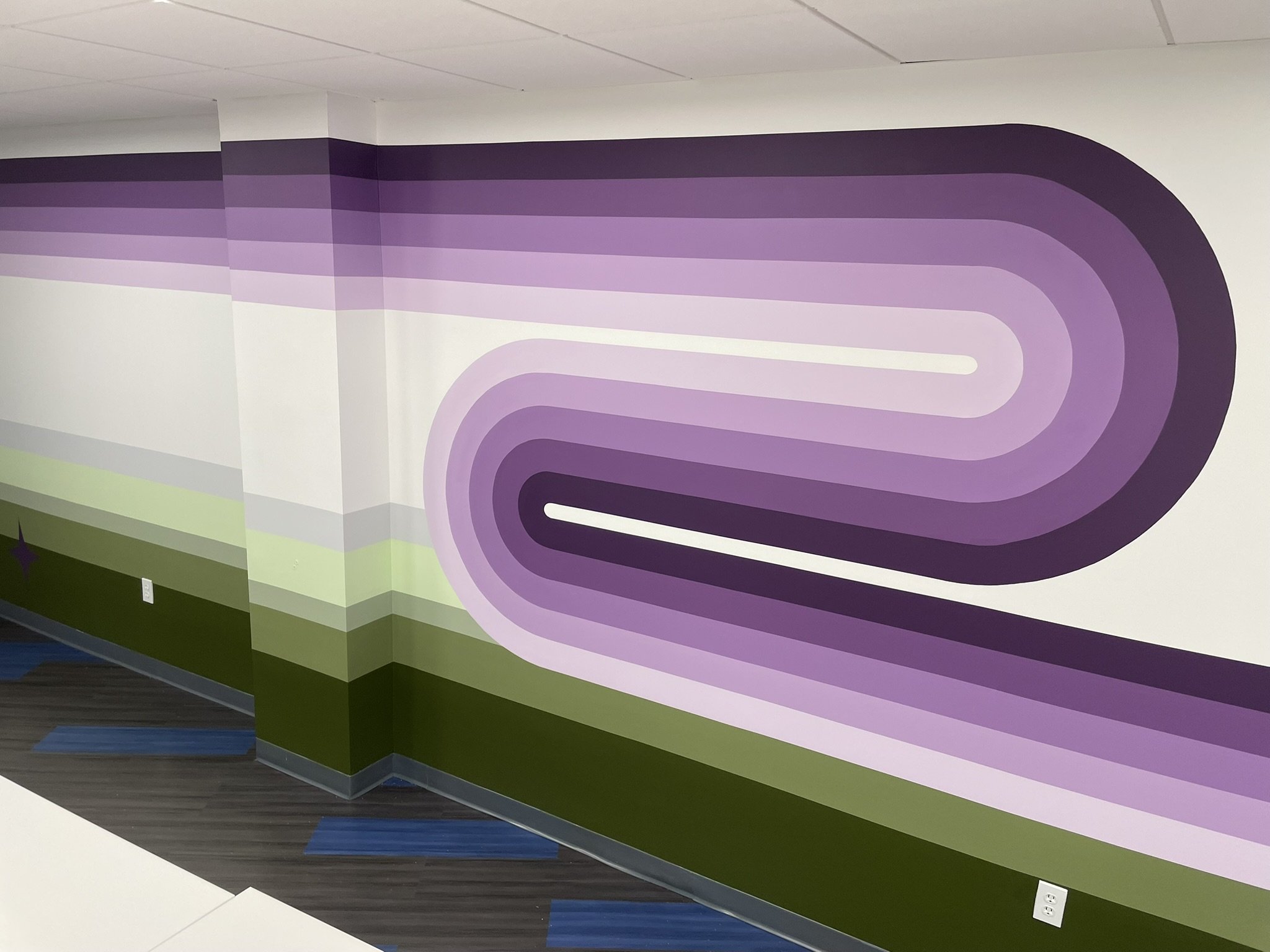 Colorful abstract mural with curved lines in shades of purple, white, and green painted on an interior wall and ceiling corner.