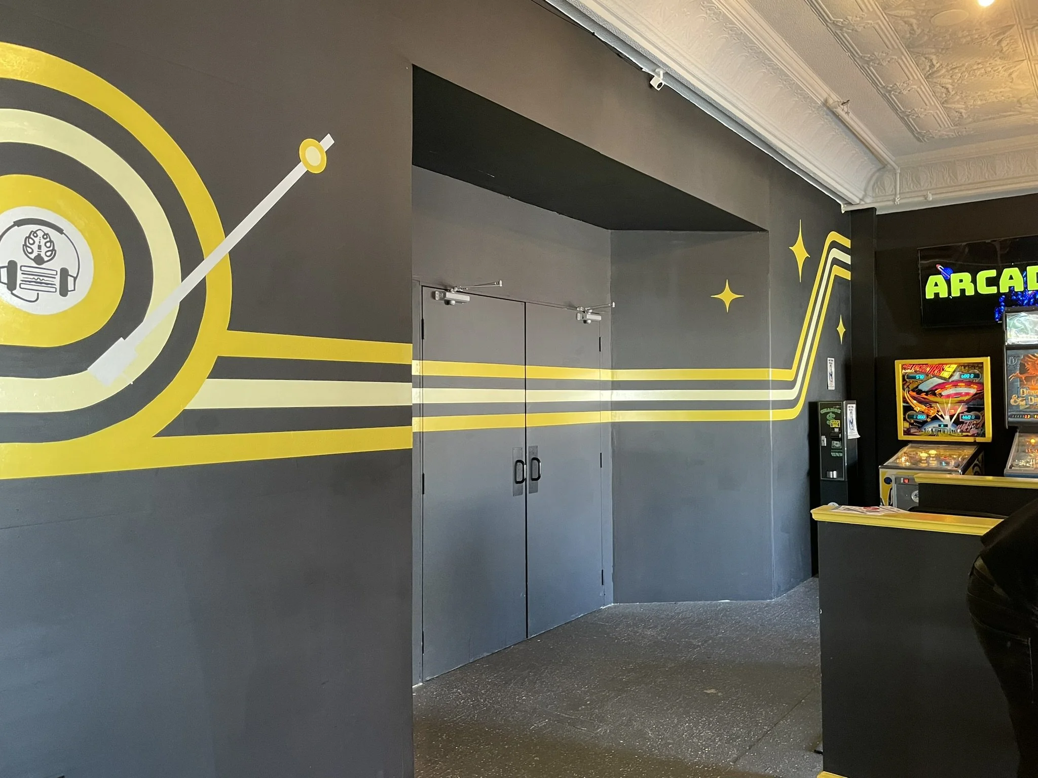 Gray wall with a yellow and black mural resembling a retro arcade game, featuring a target with a barbell icon, surrounded by lines and star-shaped highlights. There are arcade machines visible on the right side of the image.