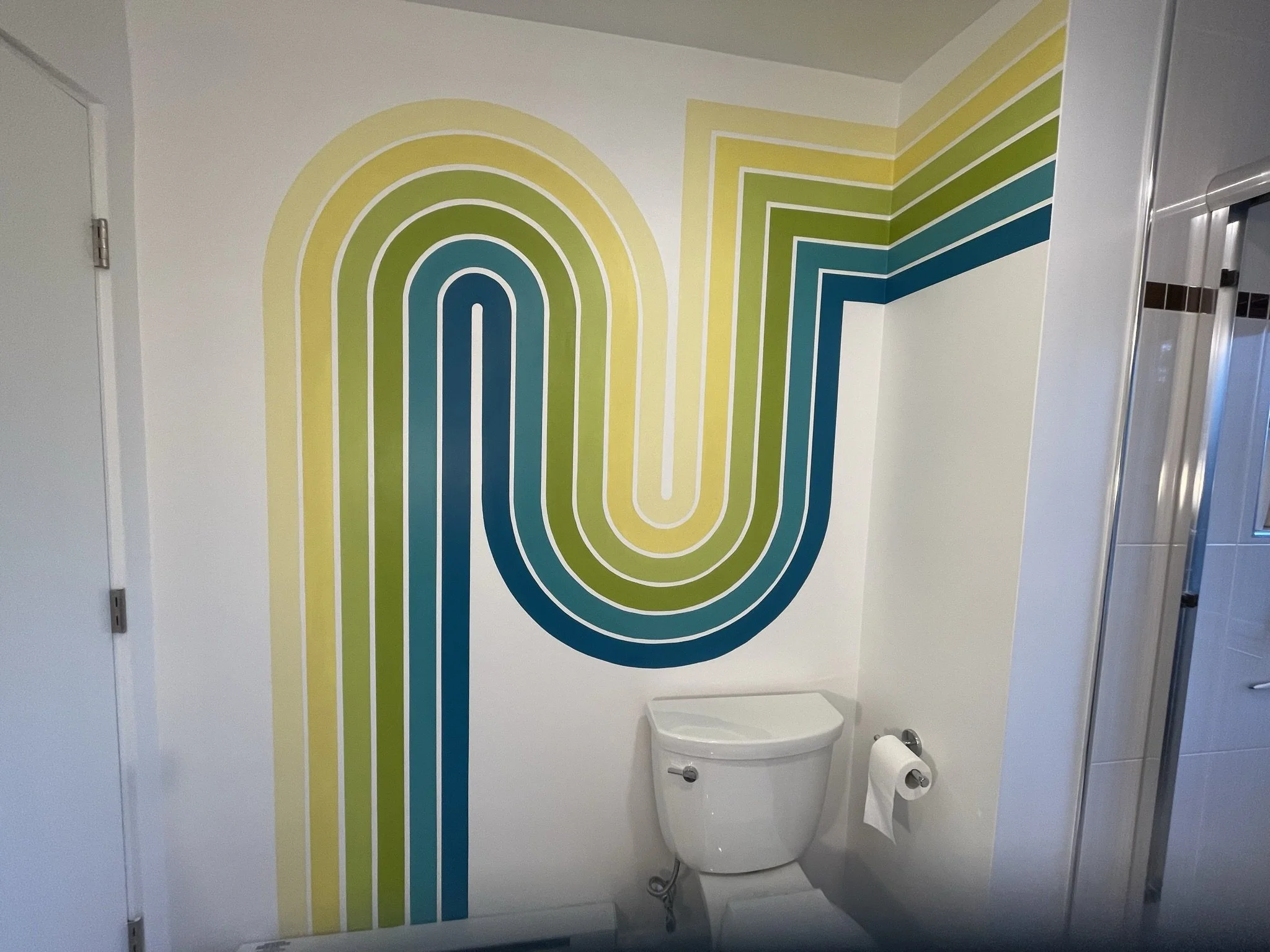 A bathroom wall decorated with a colorful, modern mural featuring curved lines in shades of yellow, green, and teal, with a white toilet and toilet paper holder underneath.