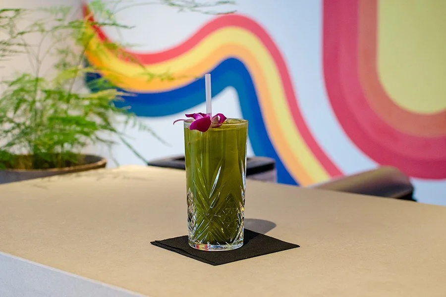 A tall glass of green beverage with a white straw and purple flower garnish on a black napkin, on a light-colored table with a colorful rainbow mural in the background and a potted plant to the left.