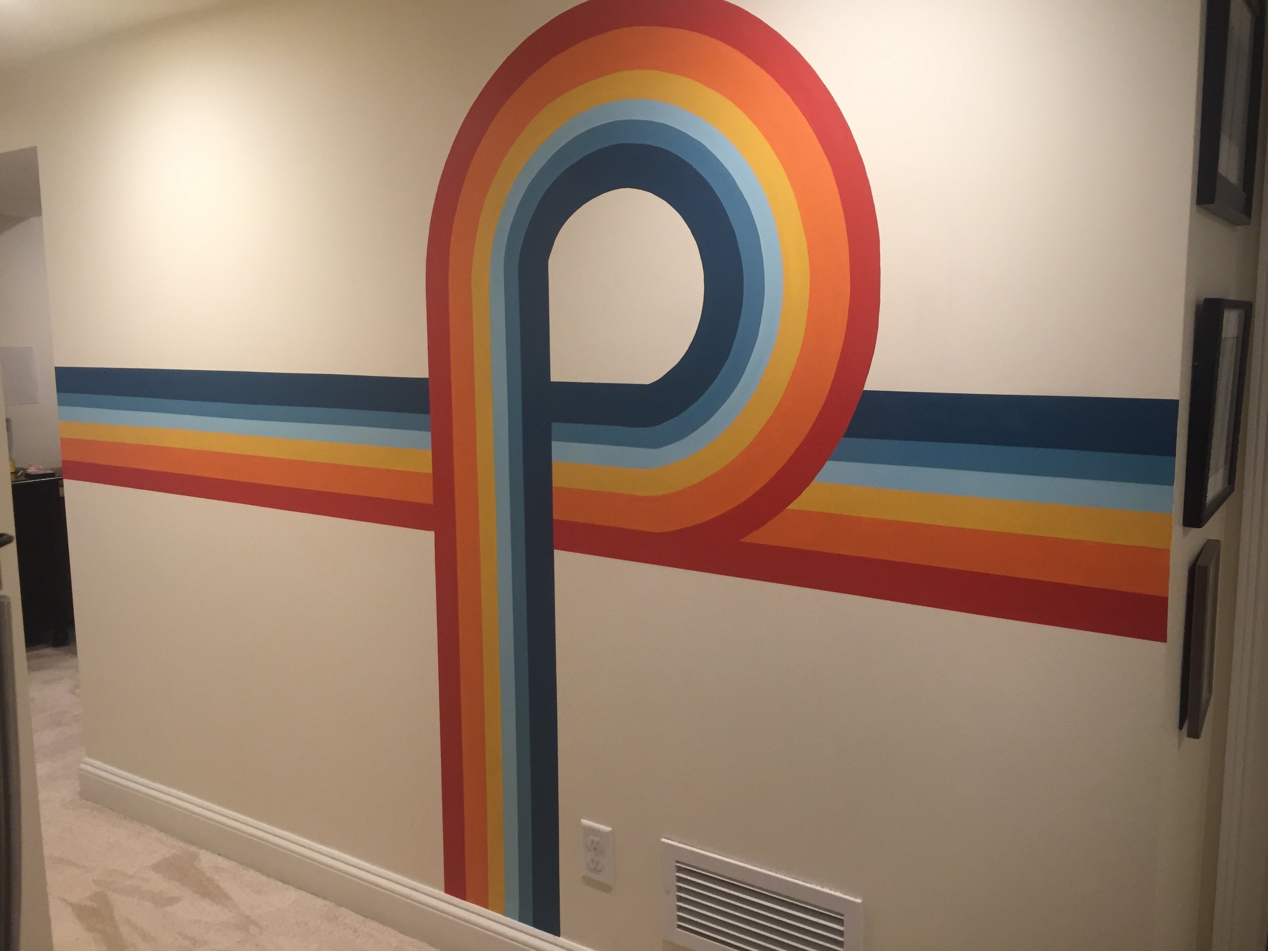 Colorful geometric wall art with a rainbow design and horizontal stripes in blue, red, orange, and yellow across a beige wall.
