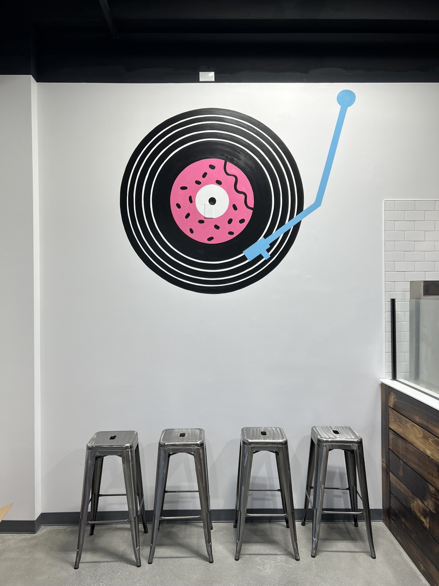 Wall art of a vinyl record with a pink donut in the center, a blue arrow pointing upward, and four metal stools in front.