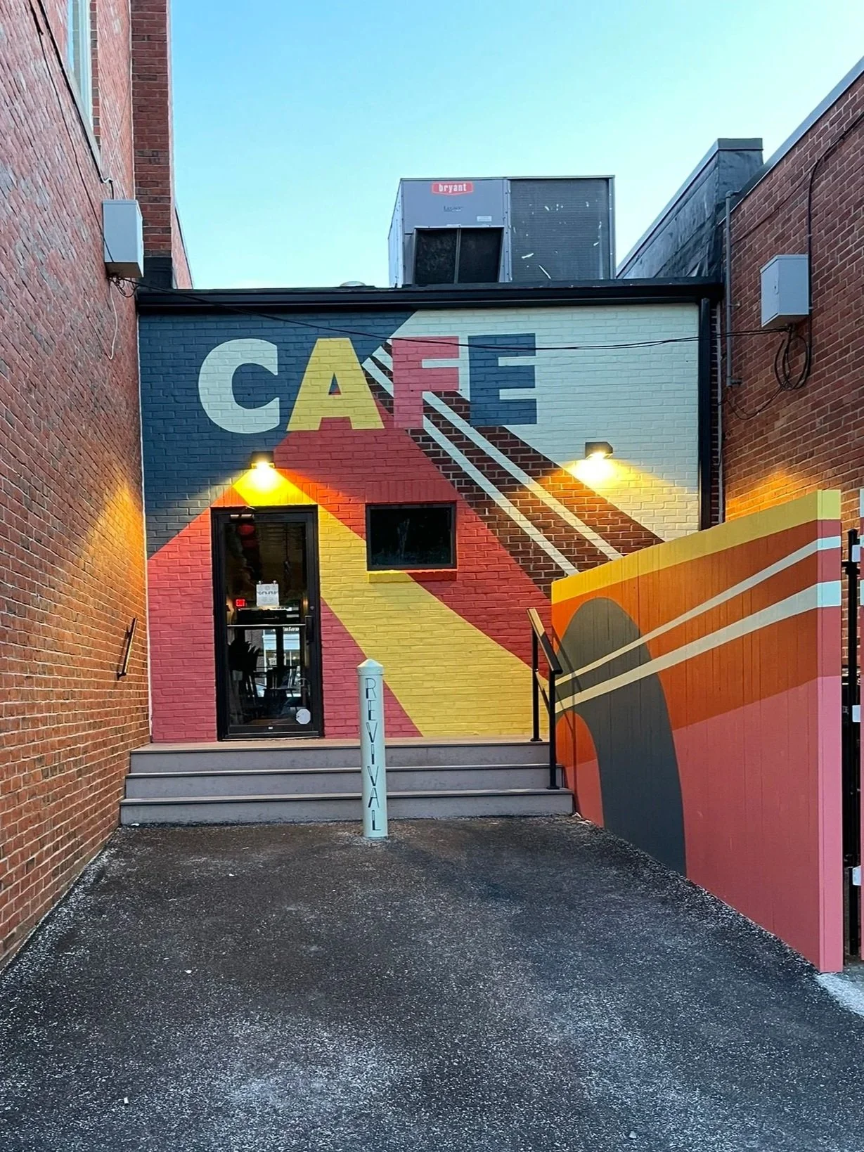 Colorful mural on the exterior wall of a cafe, spelling 'CAFE' with geometric designs and vibrant colors, with steps leading to a door and illuminated by yellow lights.